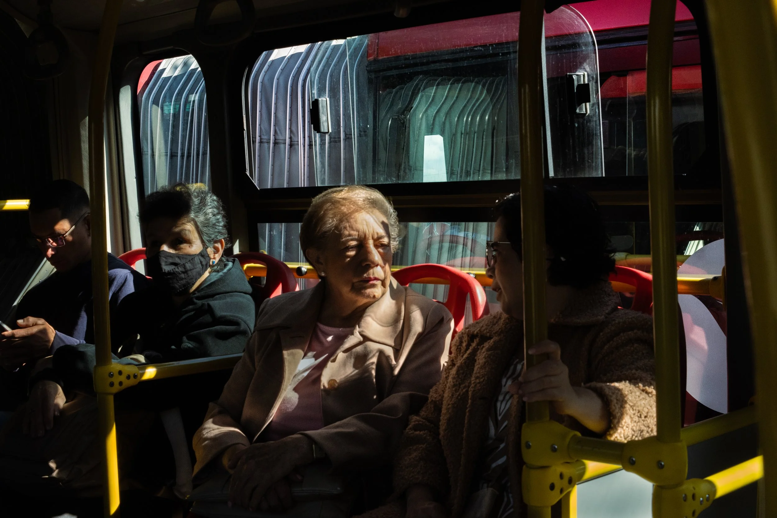 Four people sitting inside a bus with bright sunlight coming through the windows, two women are engaged in conversation, one woman is wearing a face mask, and a young man is looking down at his phone.