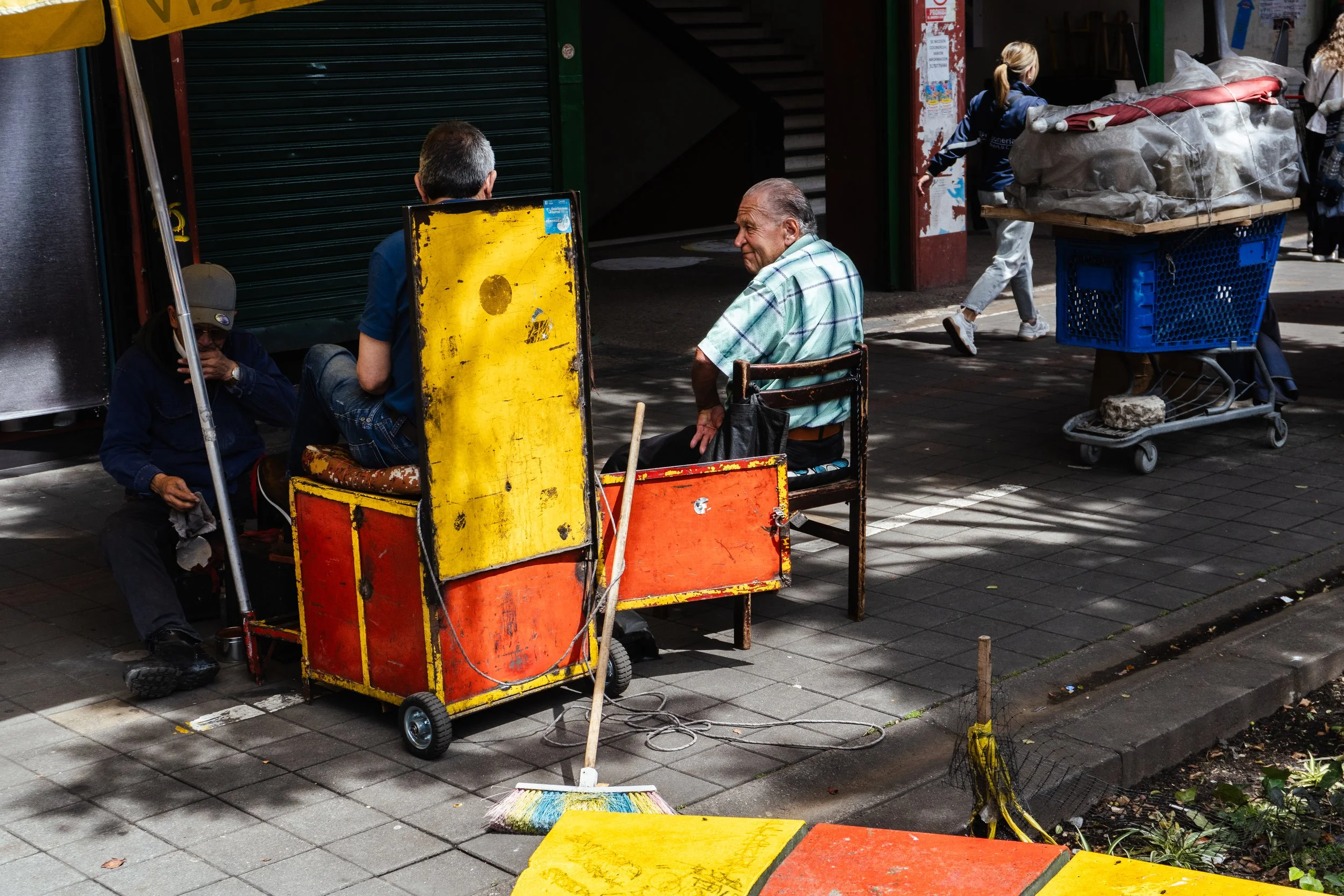Three men sitting on chairs and a bench on a city sidewalk. One man is sitting on a store's open shutter, another is shirtless wearing a striped shirt, and a third is hiding behind a yellow and red cart. There are various objects like a broom, a yell