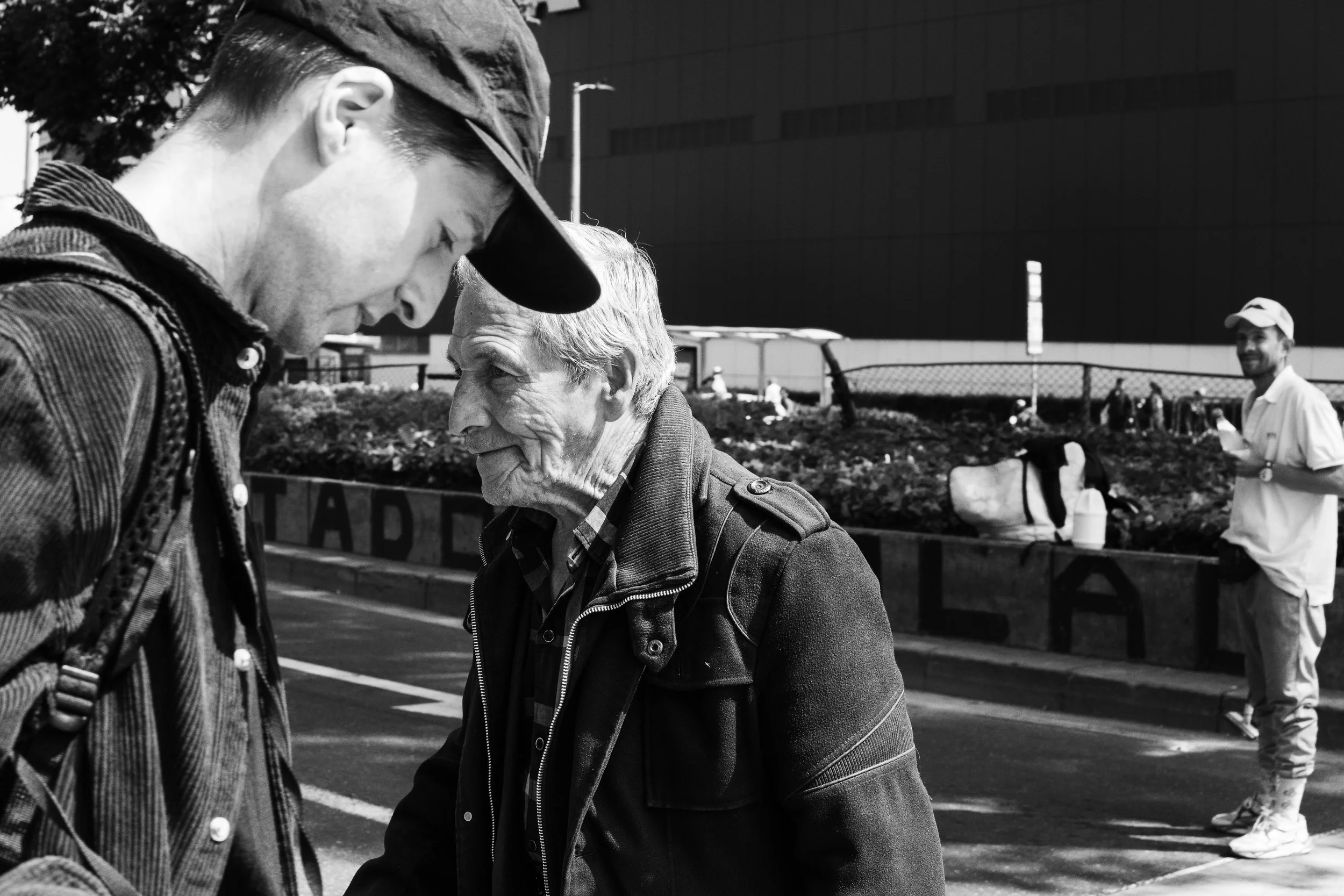 Two men are engaged in a close conversation on a city street, with a third man standing a short distance behind them, smiling and holding a smartphone.