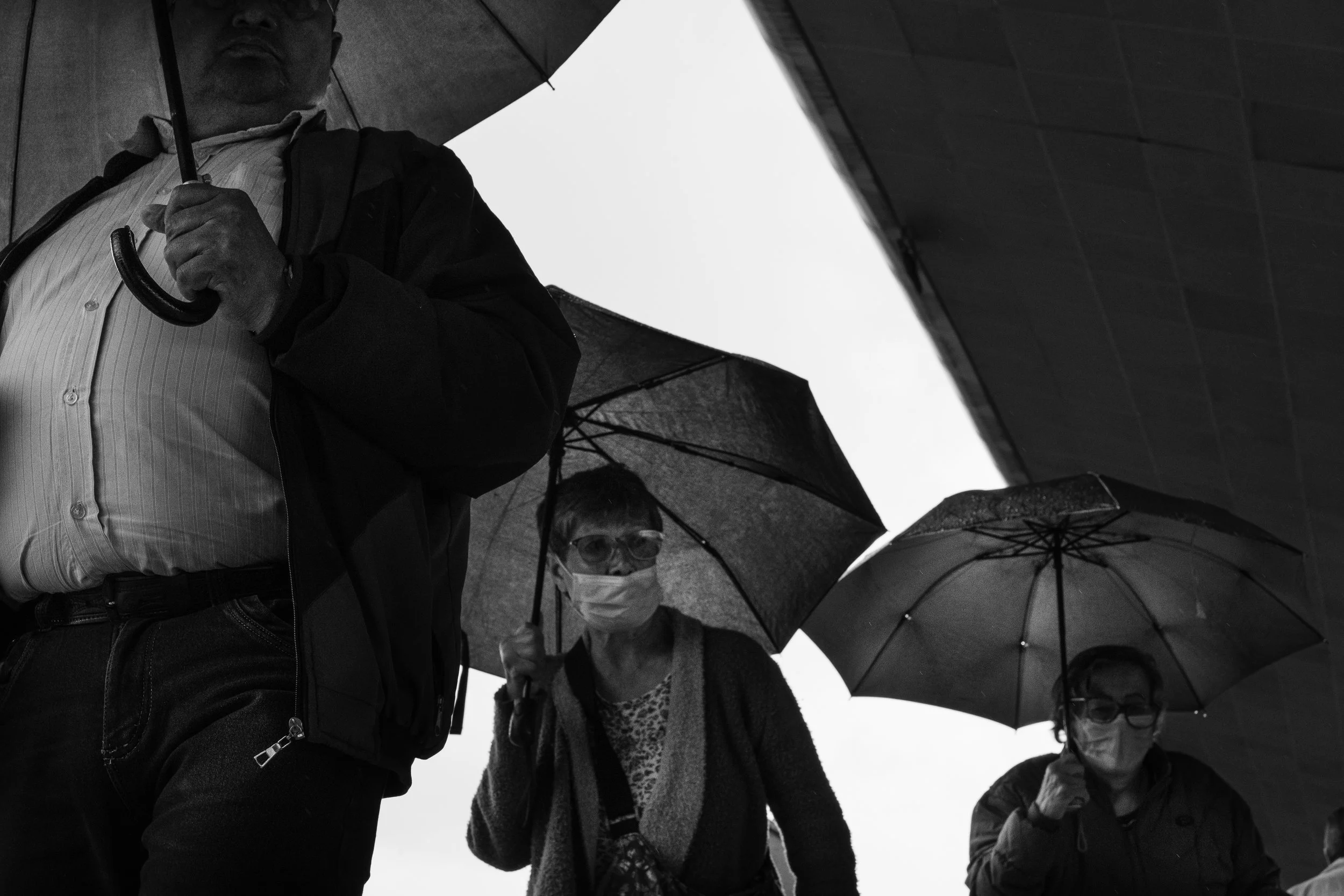 Three people holding umbrellas in rainy weather, wearing face masks, with parts of buildings visible in the background.