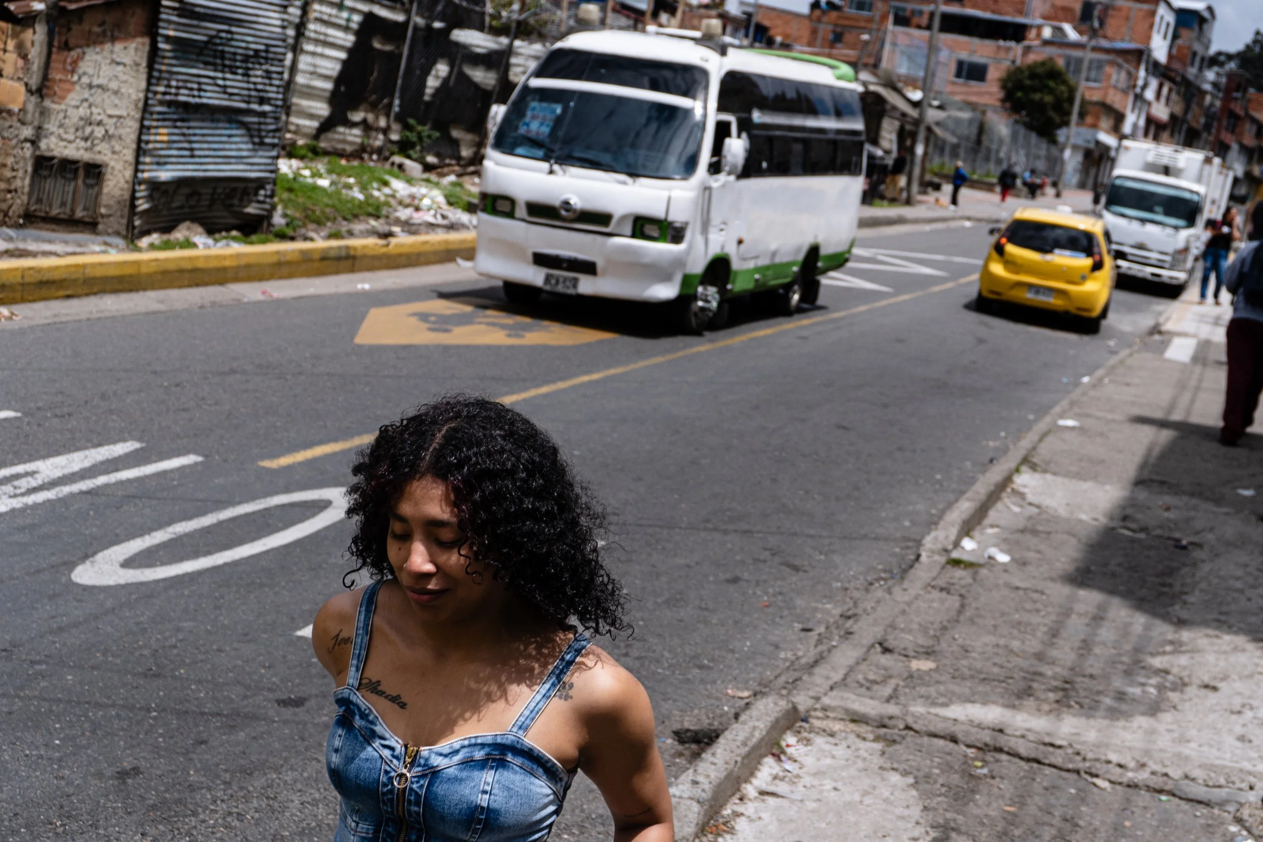 A woman with curly black hair and tattoos on her shoulders walks along a city street with vehicles in the background, including a white and green mini bus, a yellow car, and a white truck.