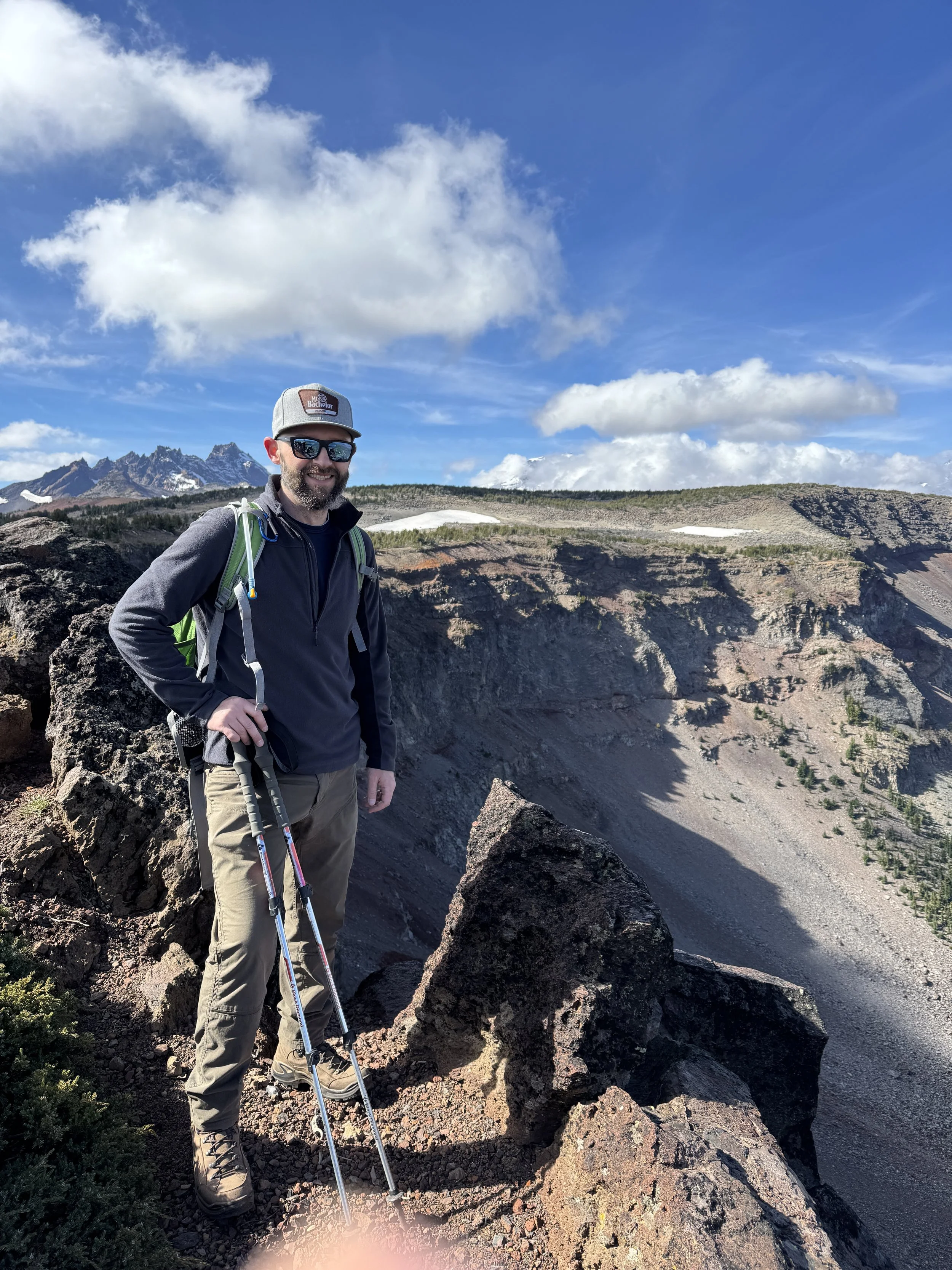 A man standing on rocky volcanic terrain, wearing hiking gear including sunglasses, a cap, and carrying hiking poles, with a mountainous landscape and blue sky with clouds in the background.