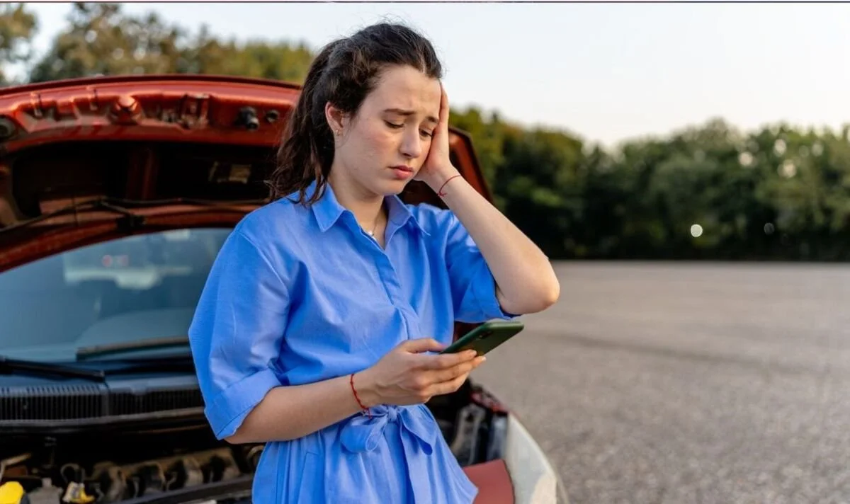 Una giovane donna con un abito blu guarda il telefono con un'espressione preoccupata, davanti a un'auto ferma sulla strada con il cofano aperto.