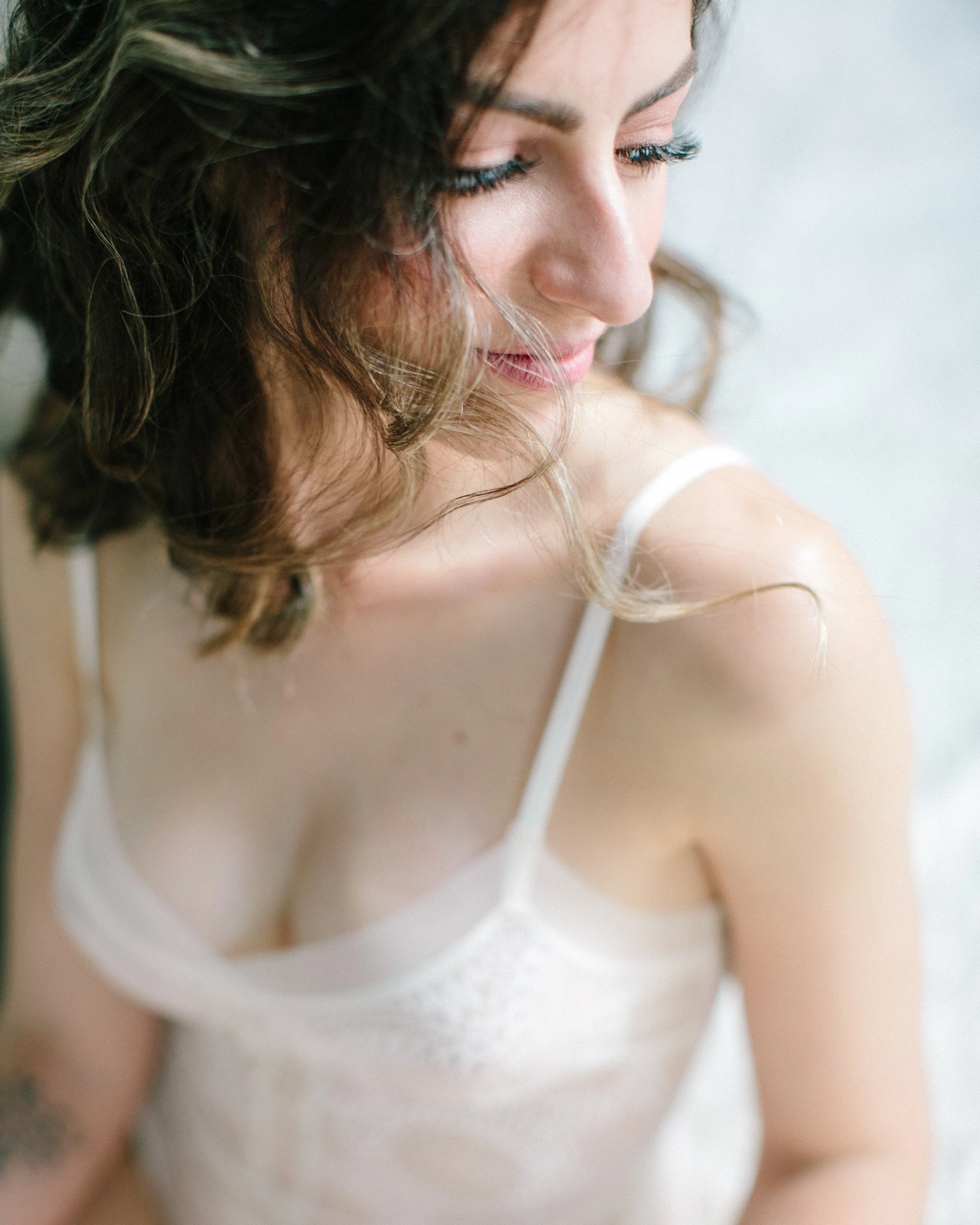 Close-up of a woman with curly brown hair and blue eyes, wearing a white bra, looking downward with a gentle expression photographed at Privée Studio in Greenville SC