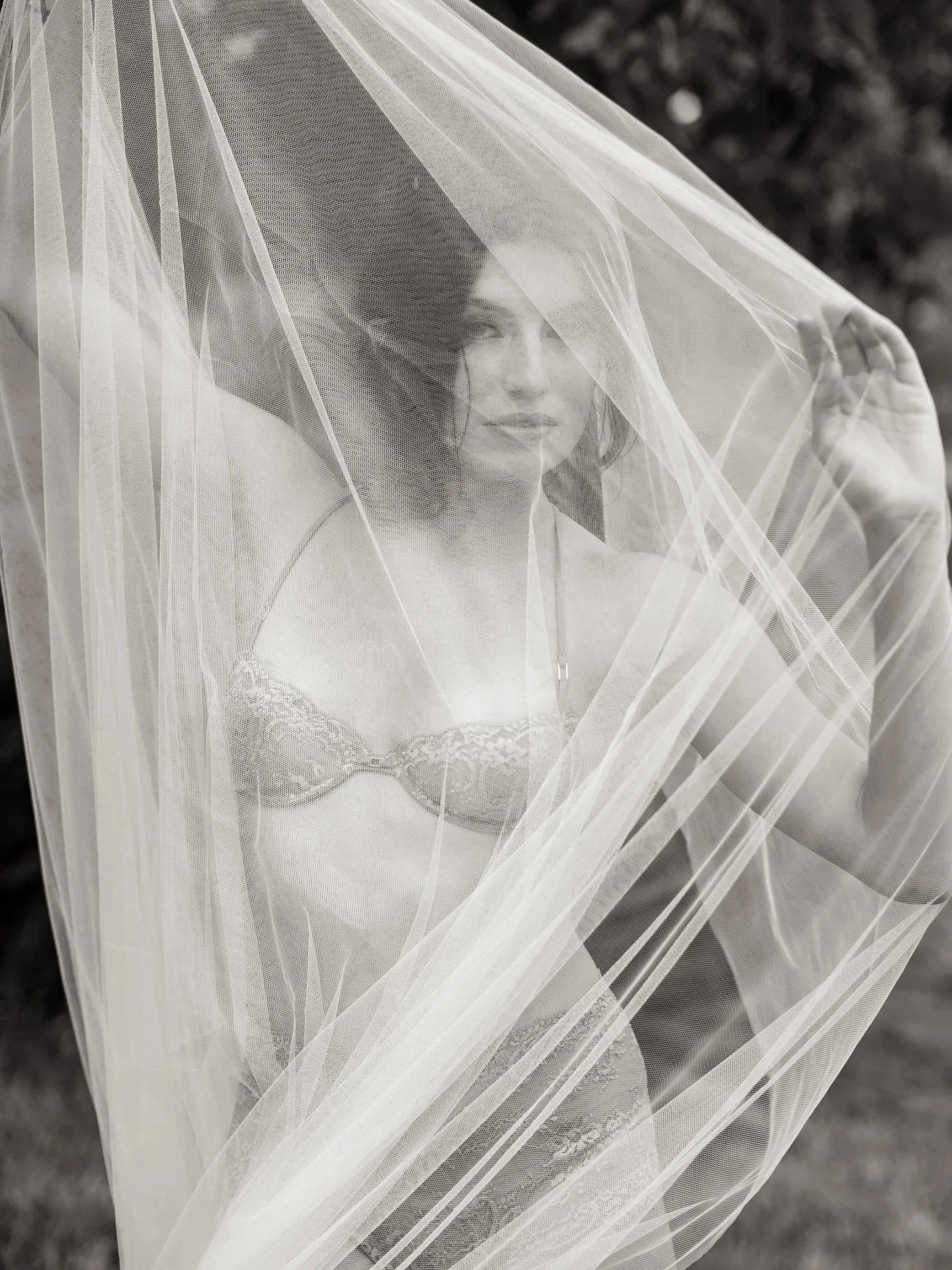 Black and white photograph of a woman wearing a lace bra, partially covered by a sheer veil, with dark hair and a serious expression photographed at Privée Studio in Greenville SC