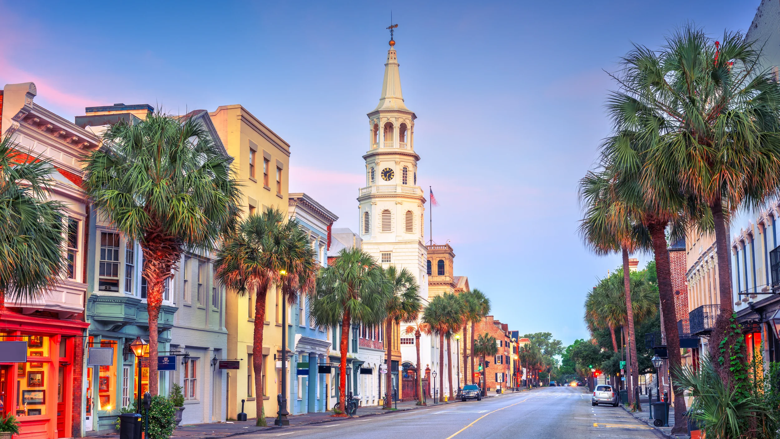A street scene with colorful historic buildings, palm trees lining the sidewalk, and a white clock tower with a pointed spire in the background, under a clear sky in the early evening.