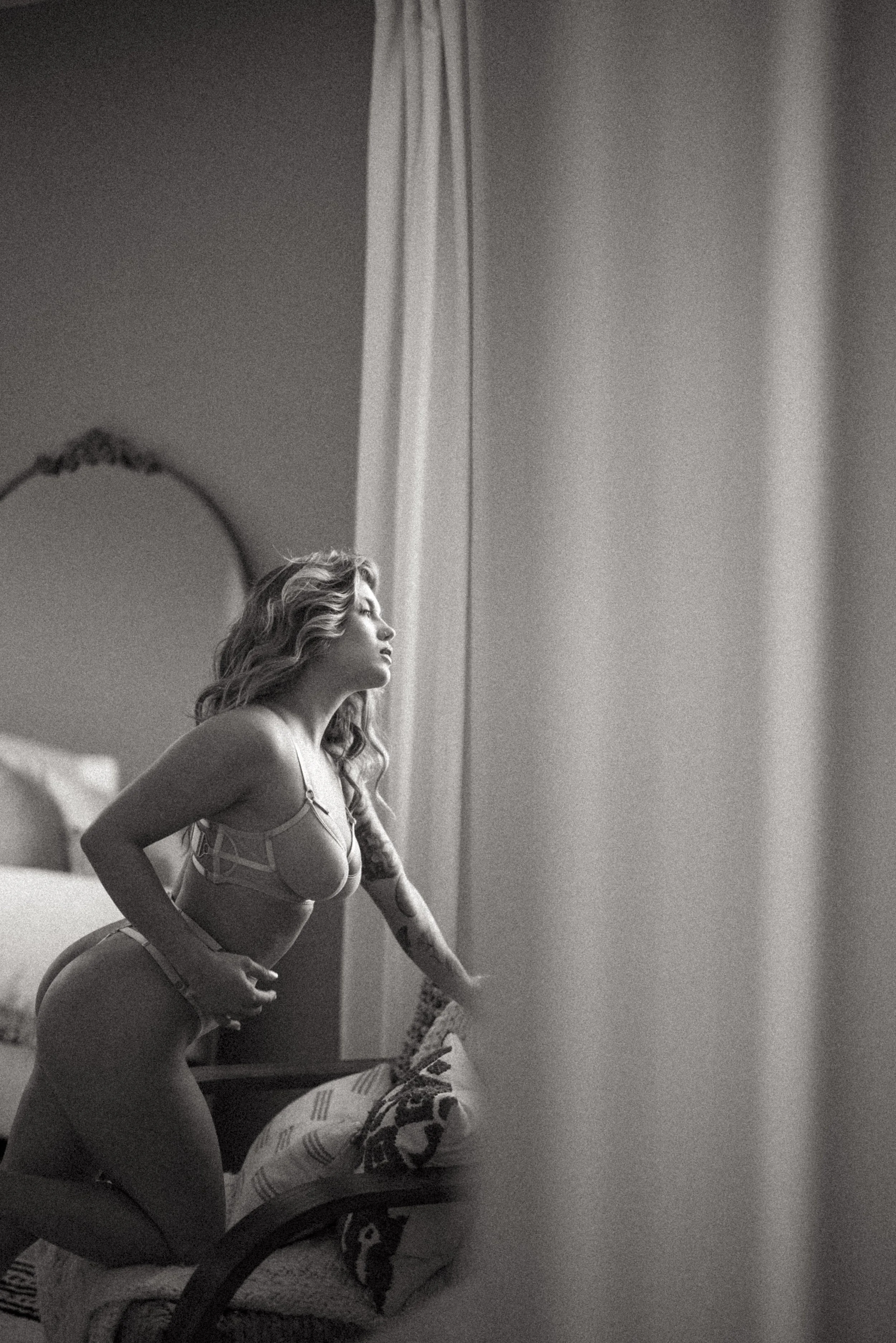 A black and white photo of a woman in lingerie sitting on a bed, resting her arm on her knee, with a mirror and curtains in the background. photographed at Privée Studio in Greenville SC