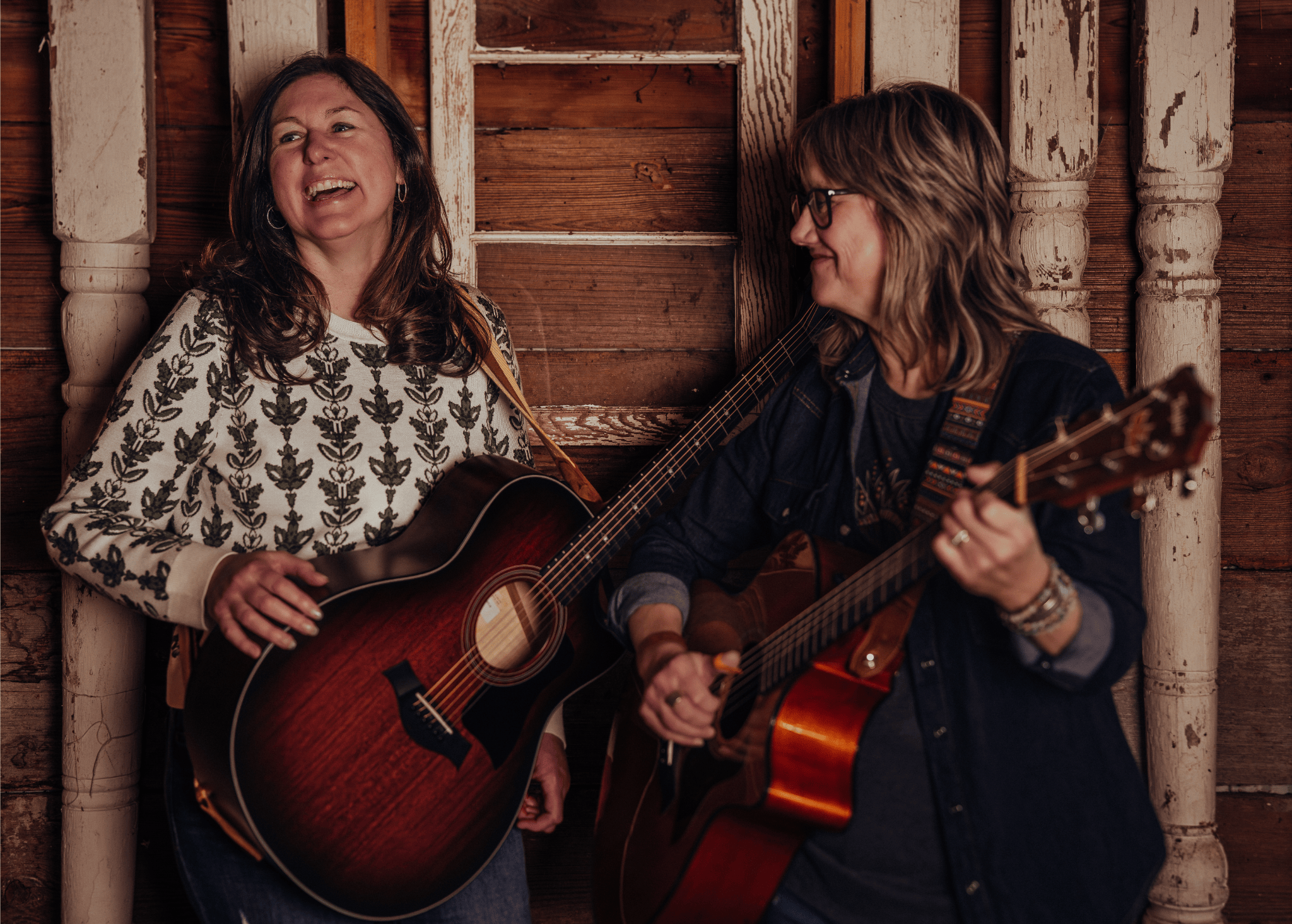Two women playing acoustic guitars and singing together in a rustic wooden room, smiling and enjoying the moment.
