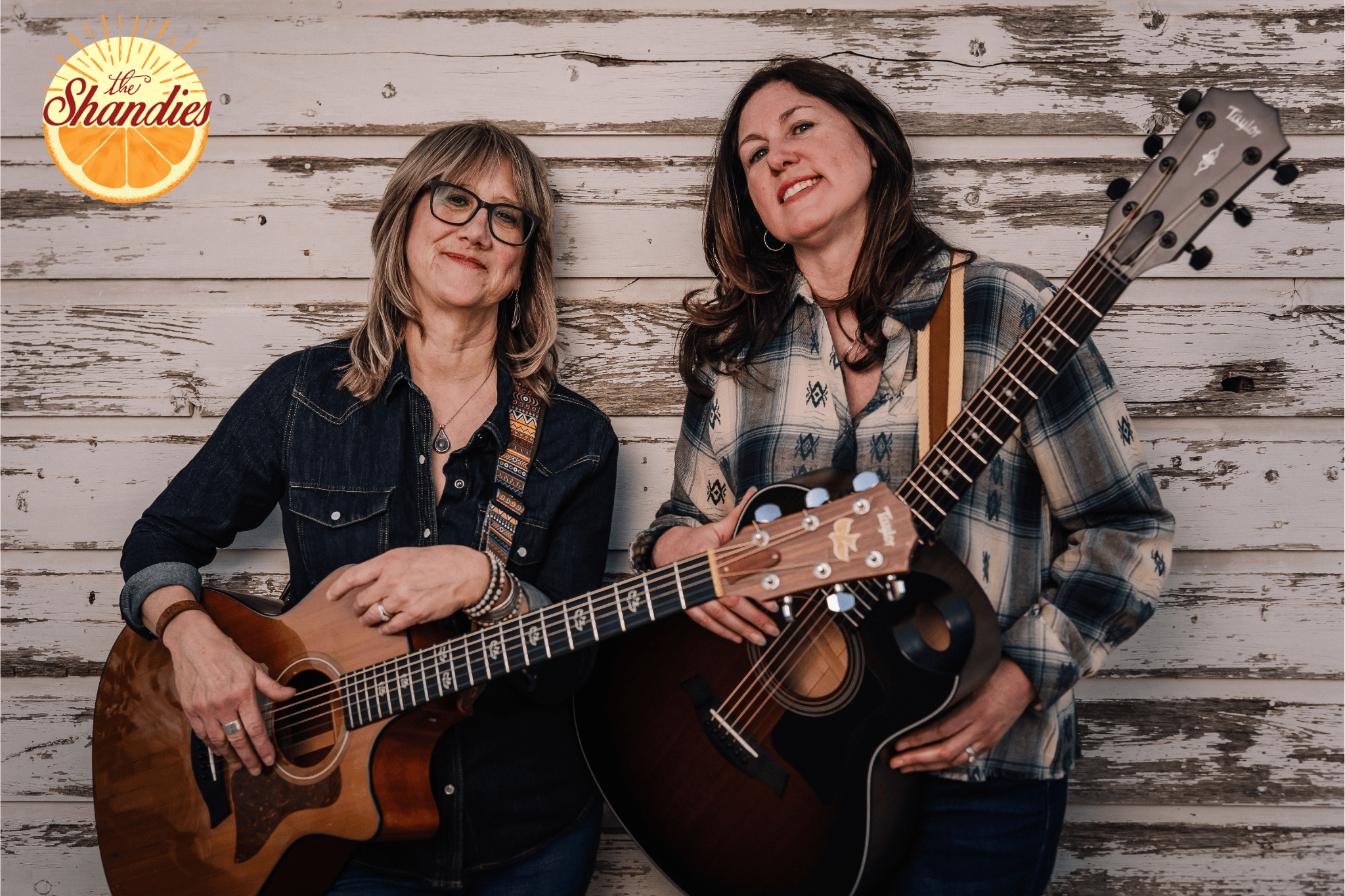 Two women with guitars standing in front of a rustic white wooden wall, smiling. One woman has glasses and shoulder-length hair, the other has long hair and is holding an acoustic guitar. A logo that says 'the Shandies' with an orange design is in the top left corner.