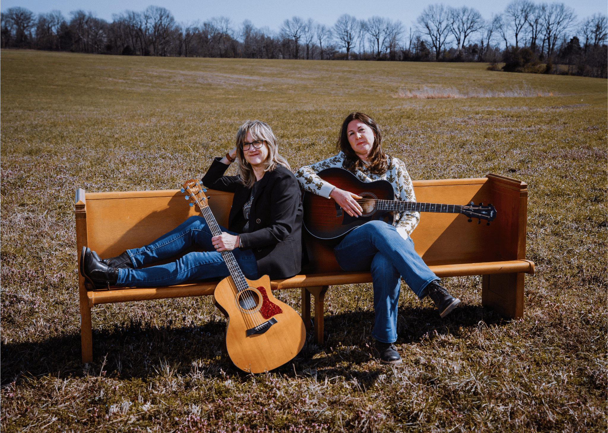 Two women sitting on a wooden bench in a field, each holding a guitar, with one sitting cross-legged and the other reclining, in a clear outdoor setting.