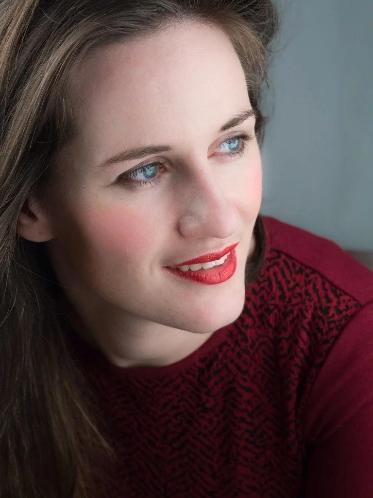 Close-up of a young woman with blue eyes, fair skin, and long brown hair, smiling slightly, wearing a red-patterned top.