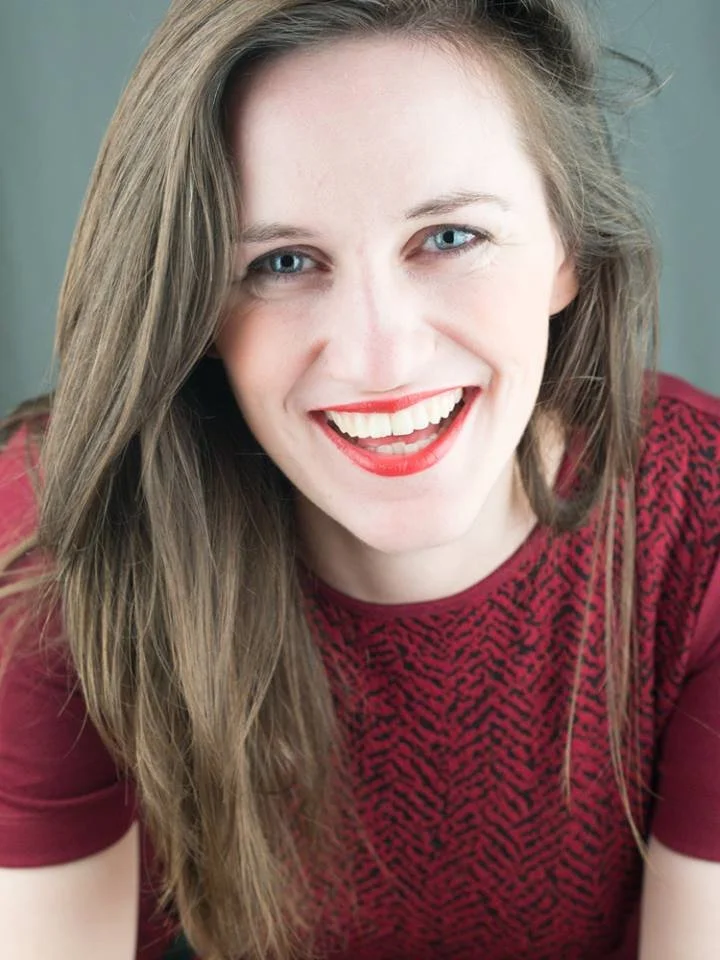 A woman with long, wavy brown hair and blue eyes, smiling with red lipstick, wearing a red patterned top.