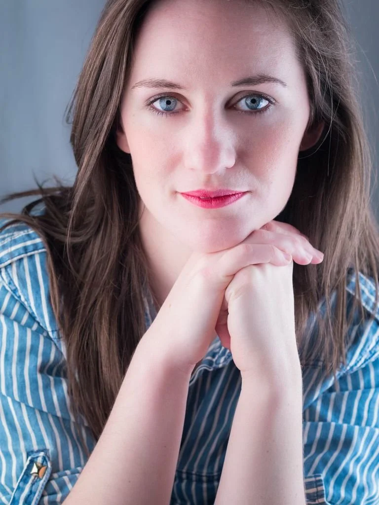 A young woman with long brown hair, blue eyes, and pink lipstick, resting her chin on her clasped hands, wearing a blue and white striped shirt, and looking directly at the camera.