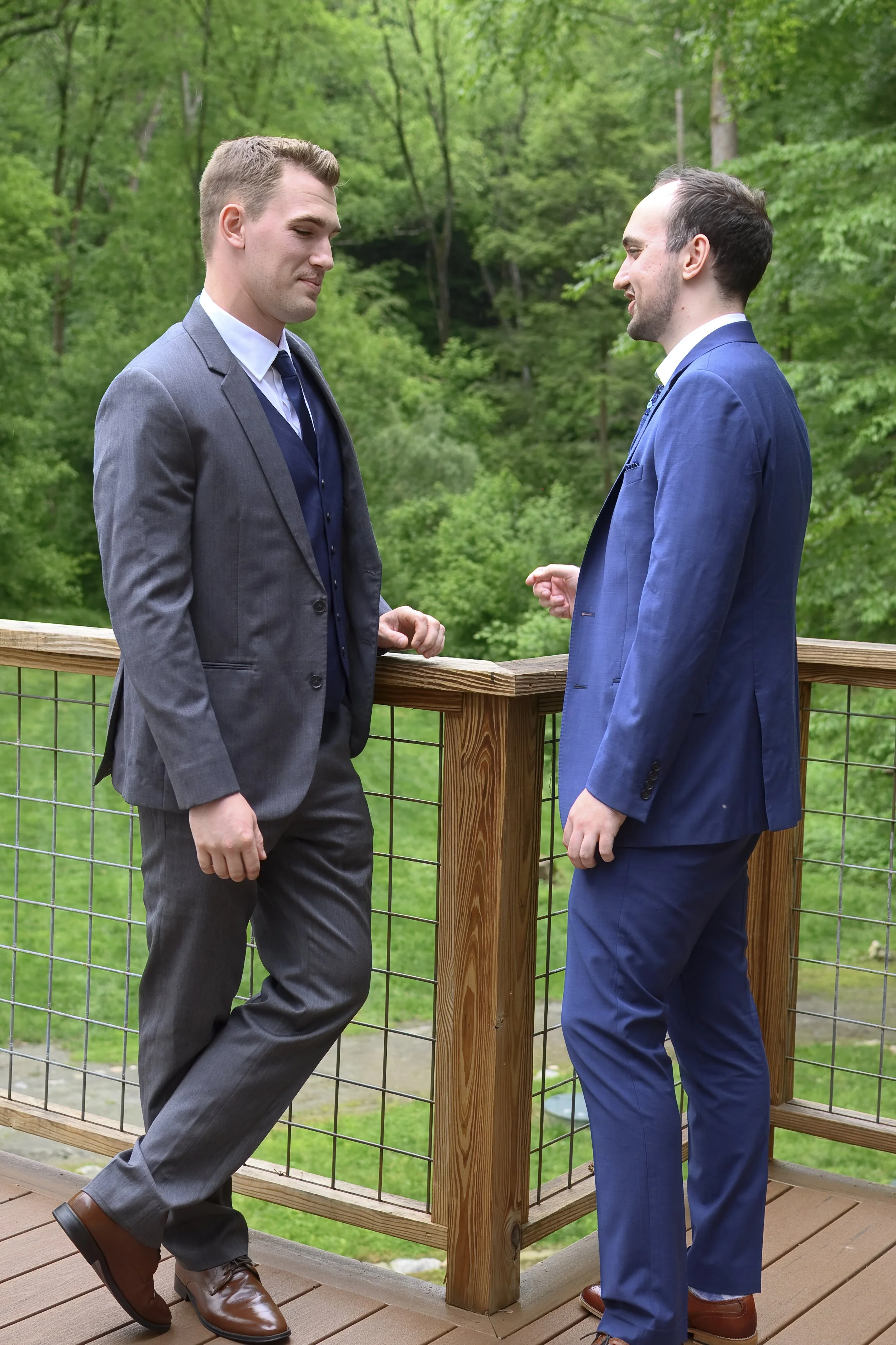 Two men in suits having a conversation outdoors on a wooden deck with a lush green forest in the background.