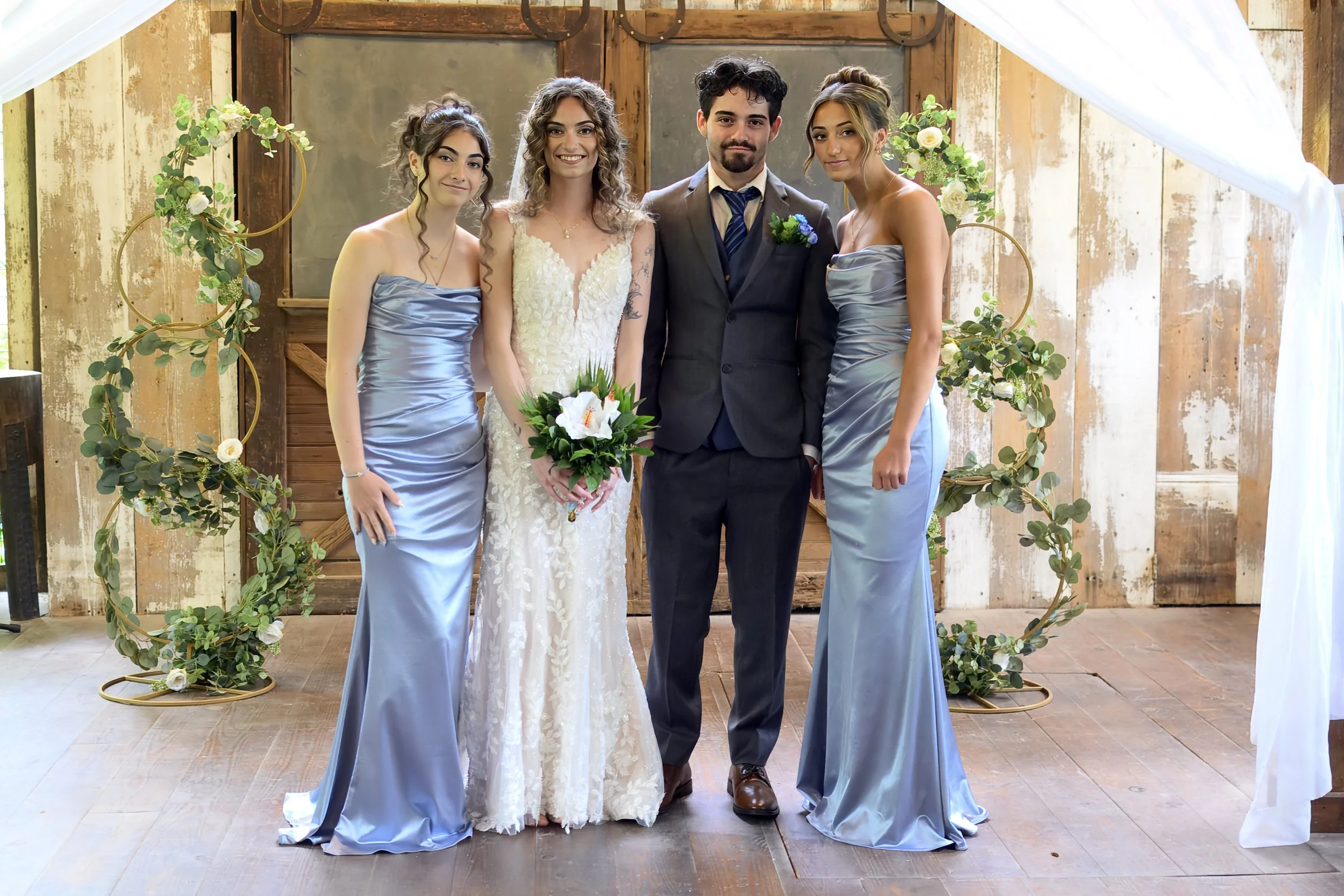A group of five people, including a bride and groom, standing together in front of a rustic wooden backdrop decorated with green leaves and white flowers, at a wedding ceremony. The bride is holding a bouquet of white flowers and wearing a lace weddi