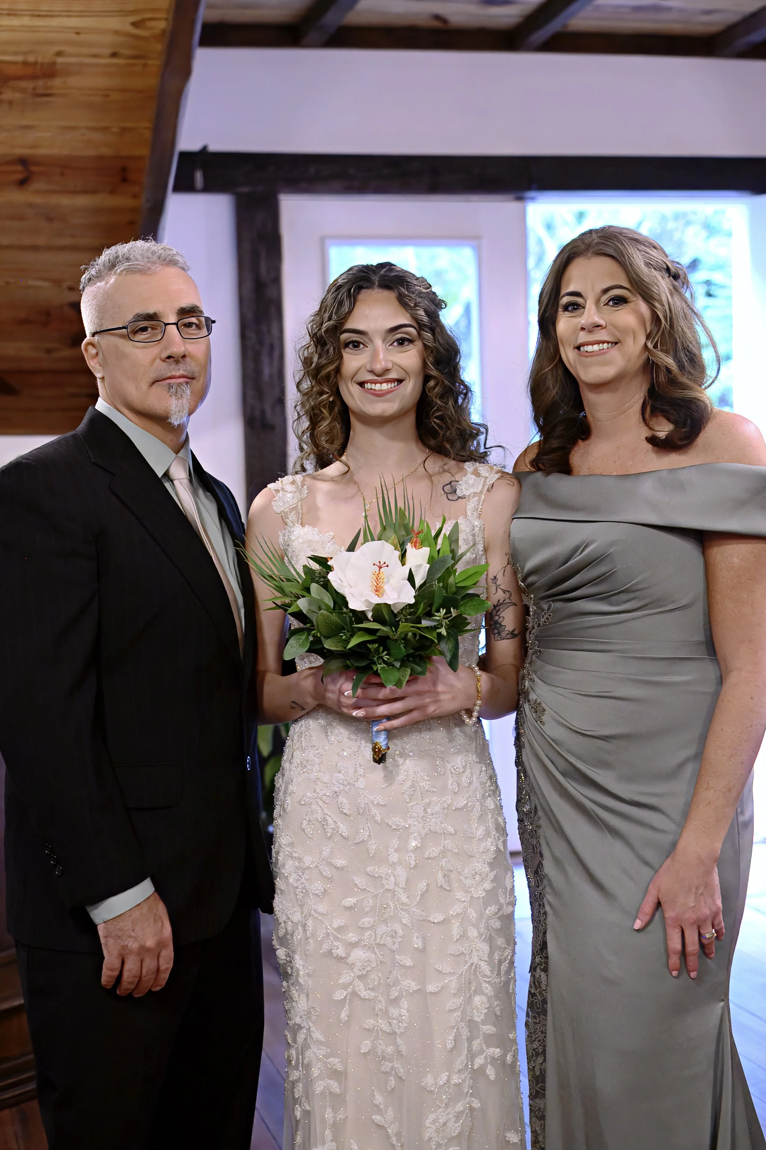 Three people standing indoors during a wedding. A man in a black suit and glasses on the left, a woman in a white lace wedding dress holding a bouquet of flowers in the middle, and a woman in a gray dress on the right.