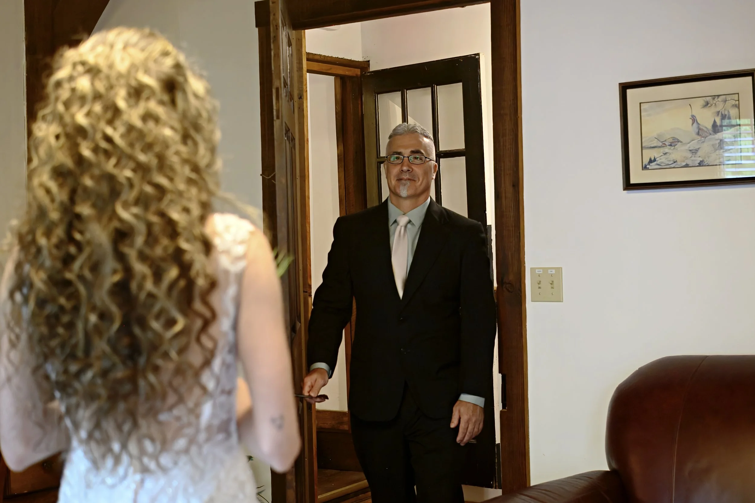 A man in a black suit with glasses and a tie is standing in a doorway, looking at a woman with curly blonde hair, who is partially visible in the foreground. The room has wooden trim and a framed picture of birds on the wall.