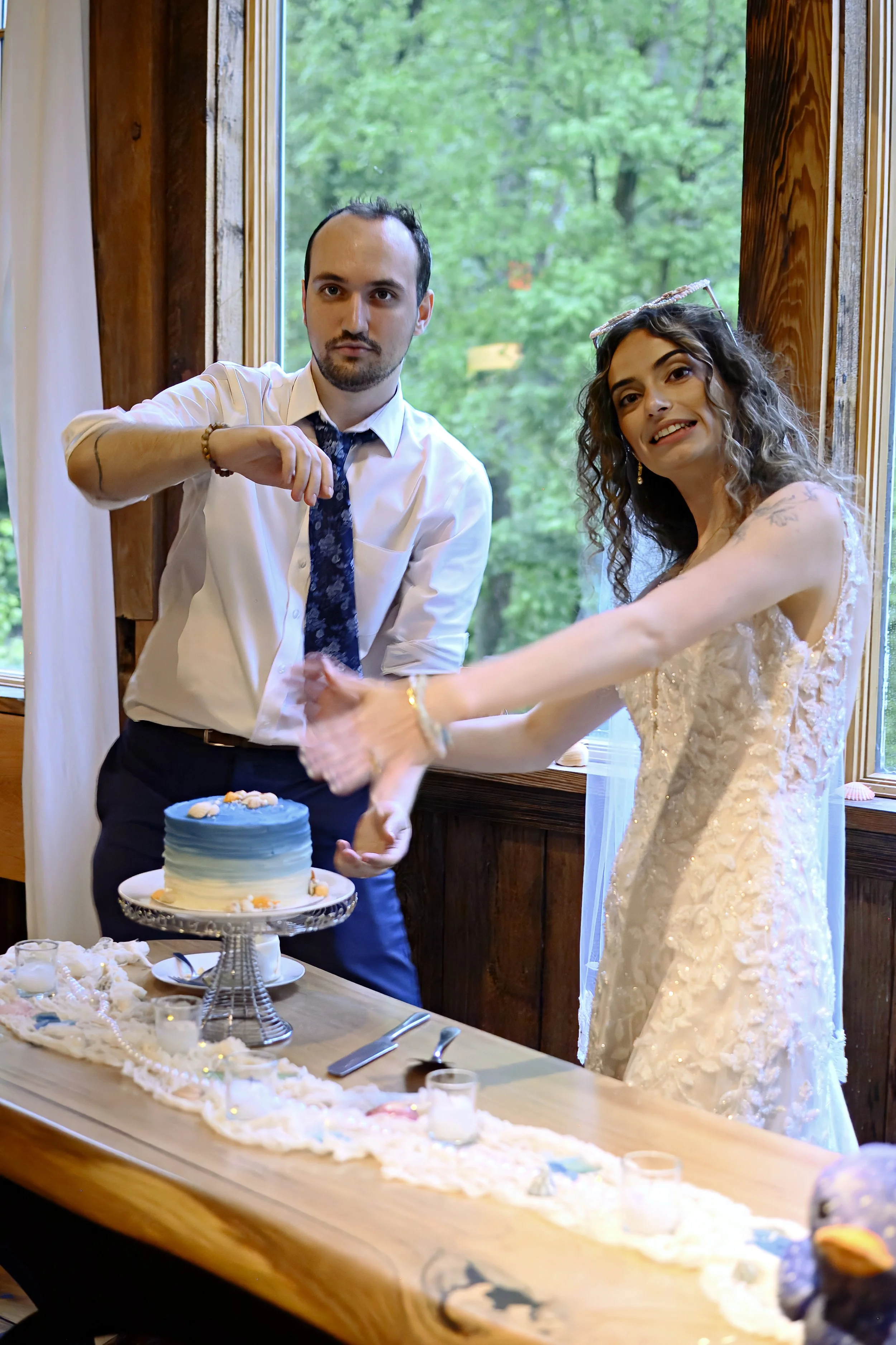 A man in a white shirt and a woman in a wedding dress standing behind a decorated table with a cake, cutting the cake together during a celebration, with a window showing green trees outside.