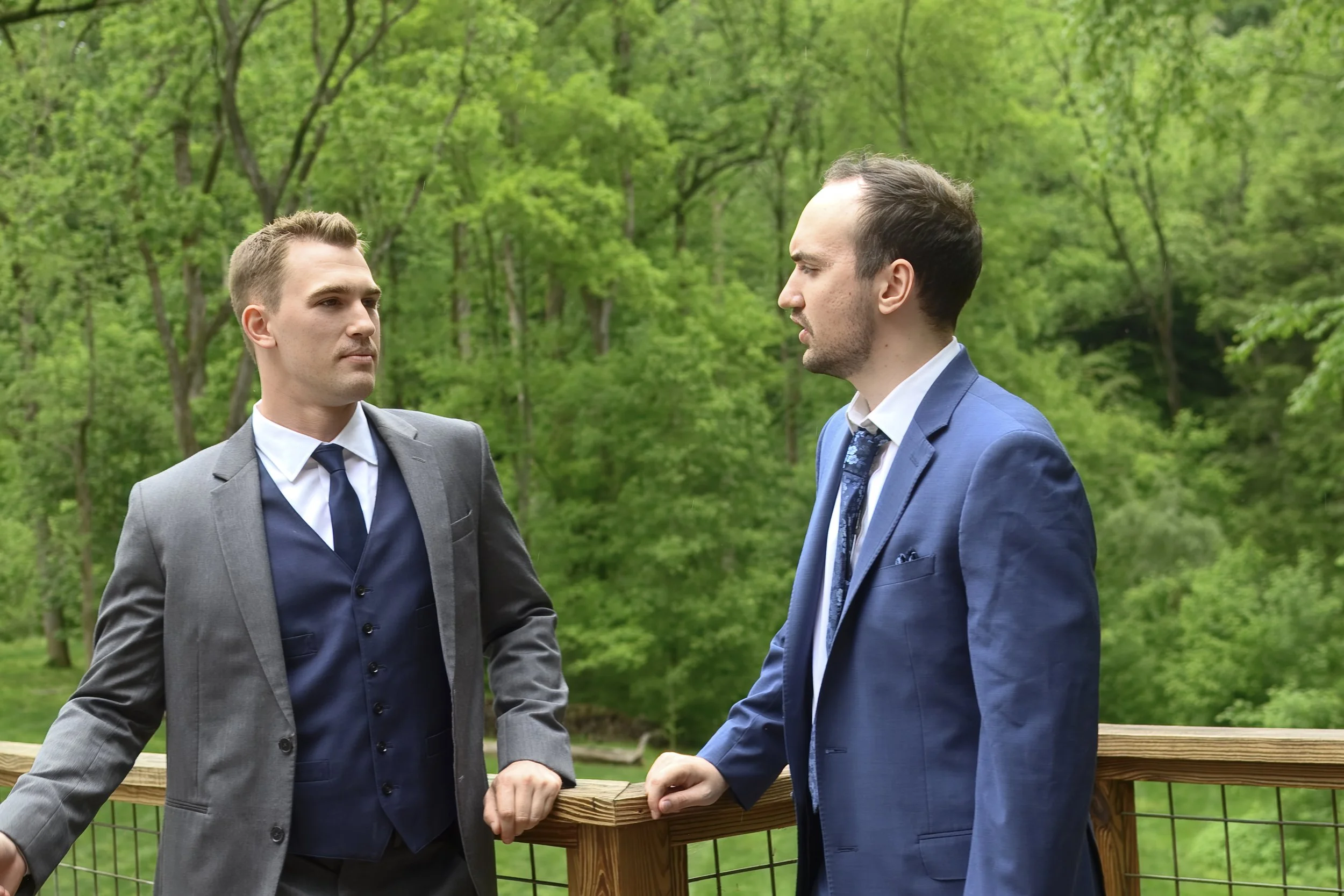 Two men in suits having a serious conversation outdoors, standing by a wooden railing with a green wooded background.