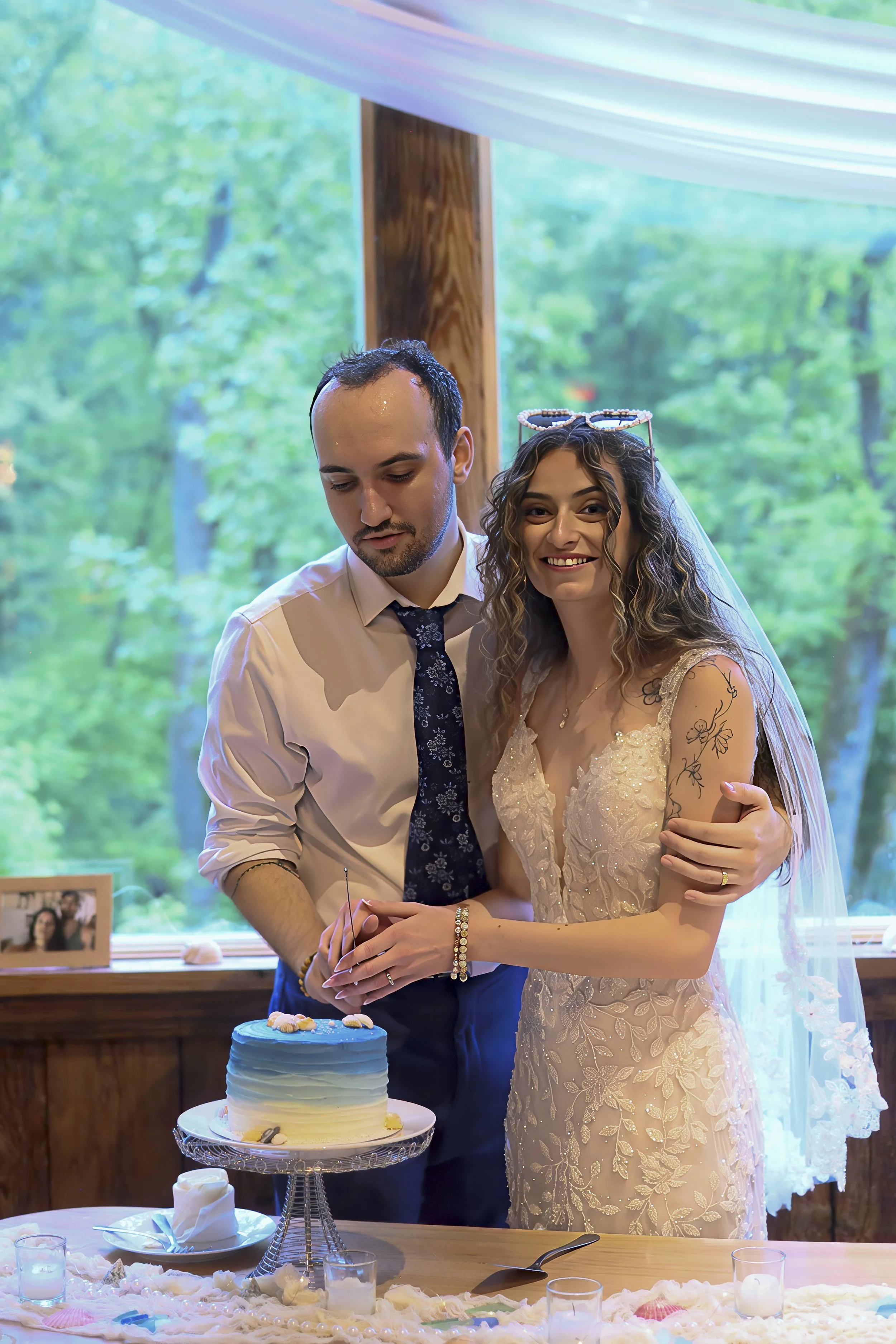 A bride and groom at their wedding reception, cutting a cake together. The bride is smiling and has curly hair, wearing a lace wedding dress with a veil. The groom is looking down, wearing a white shirt and a dark tie. There are decorations on the ta