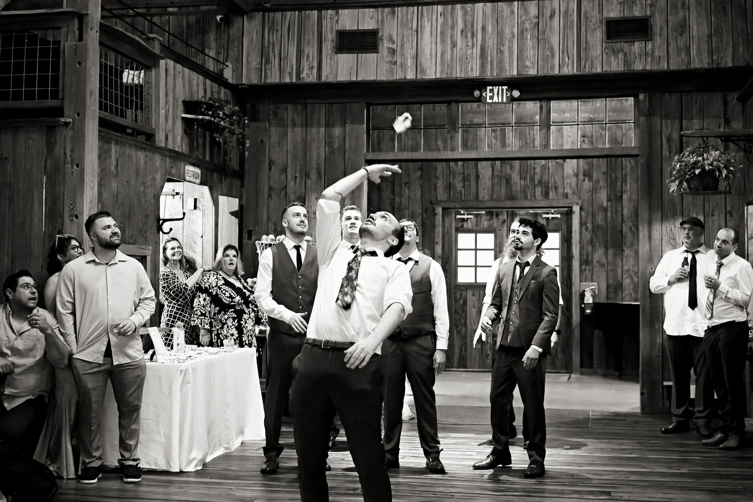 Group of people gathering indoors with wooden walls, some watching a man tossing a paper napkin into the air, others smiling and observing, in a rustic setting.