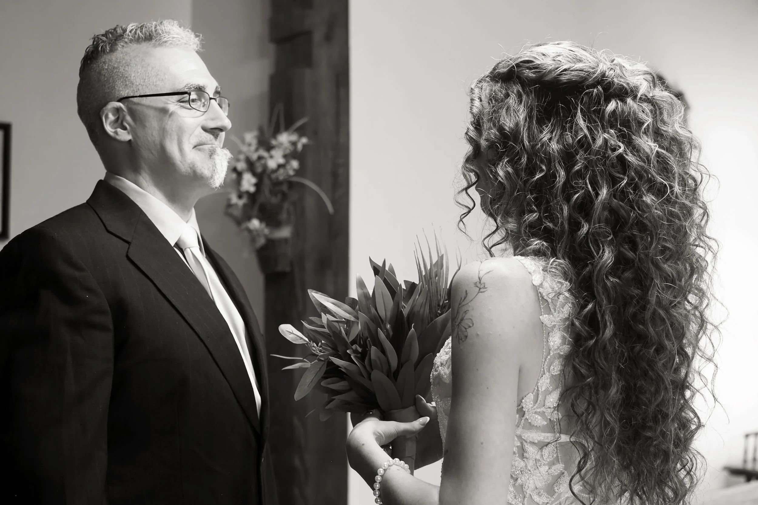 A man in a tuxedo with glasses and a woman with long curly hair in a wedding dress exchanging wedding rings.