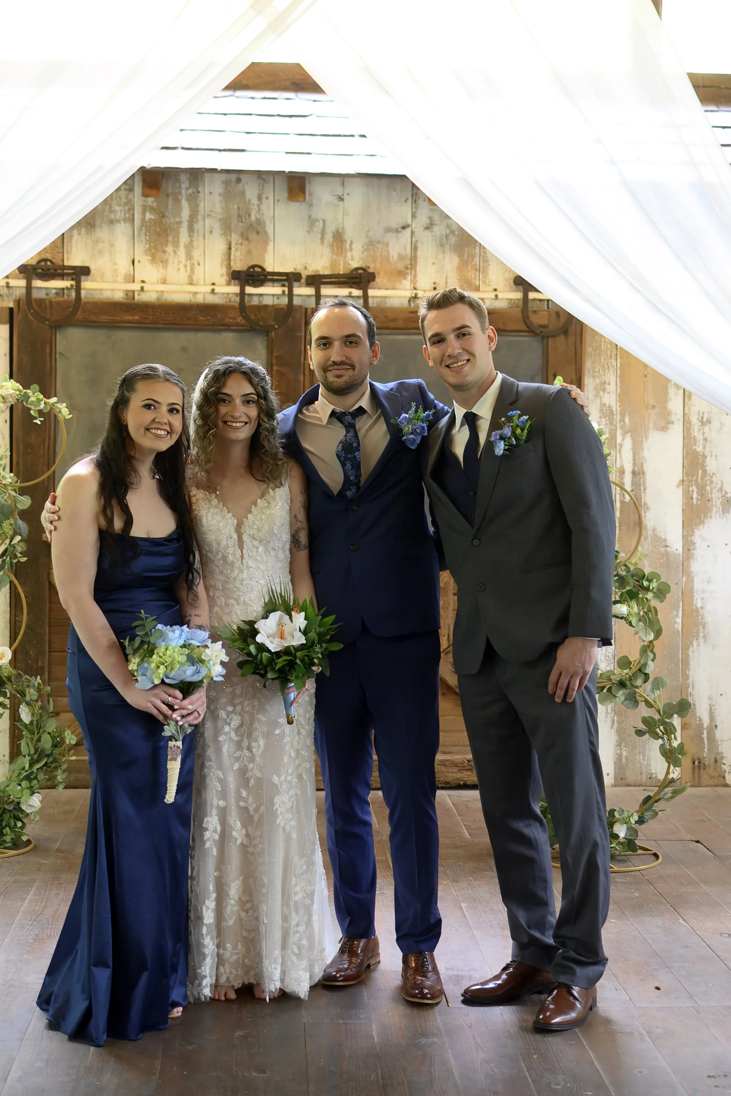 Group of four people at a wedding, three men and two women, dressed formally standing together indoors with rustic wood background and greenery.