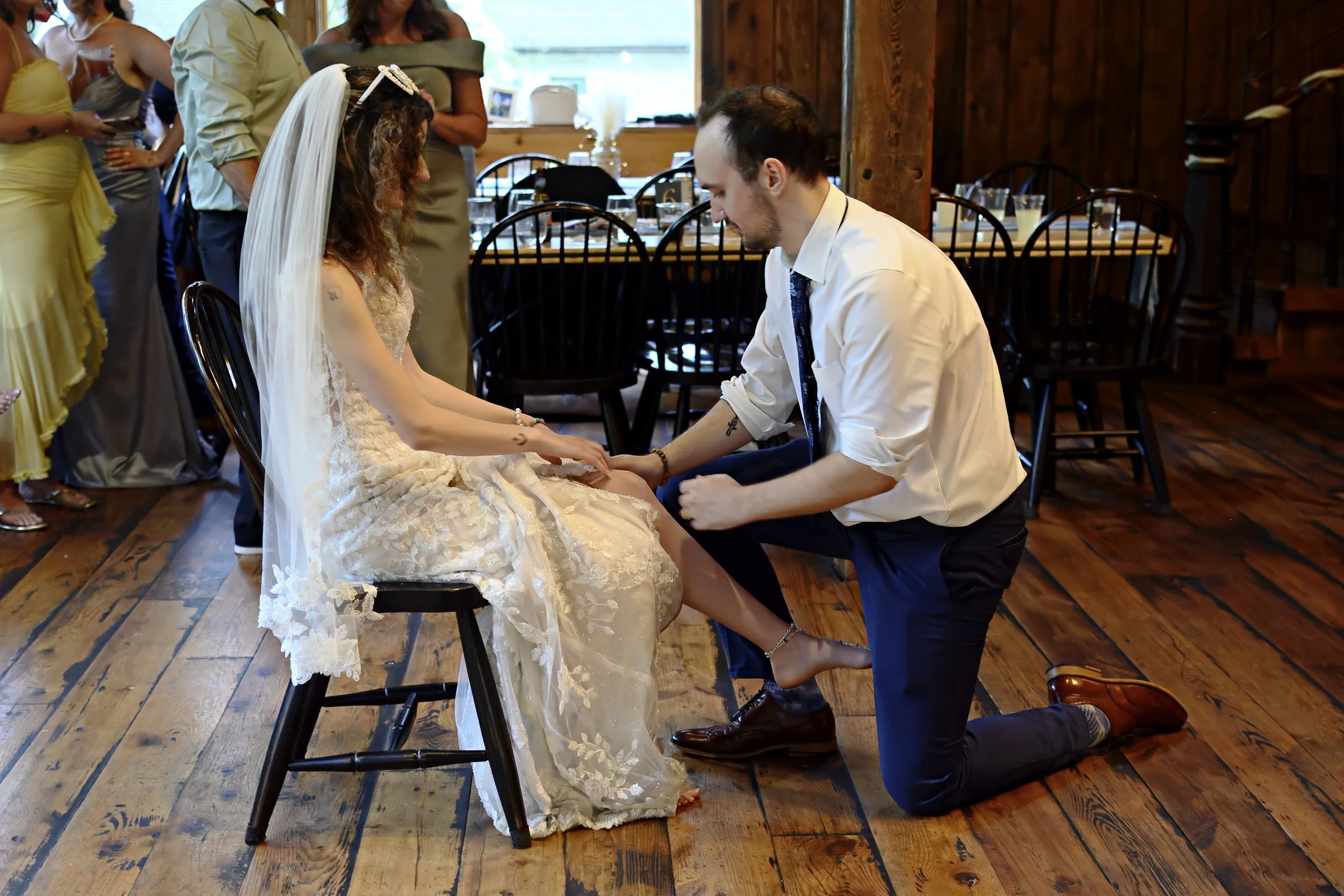 A man kneels before a seated woman in a wedding dress, holding her hands and appearing to be proposing or exchanging vows, inside a rustic wooden venue with several guests watching in the background.