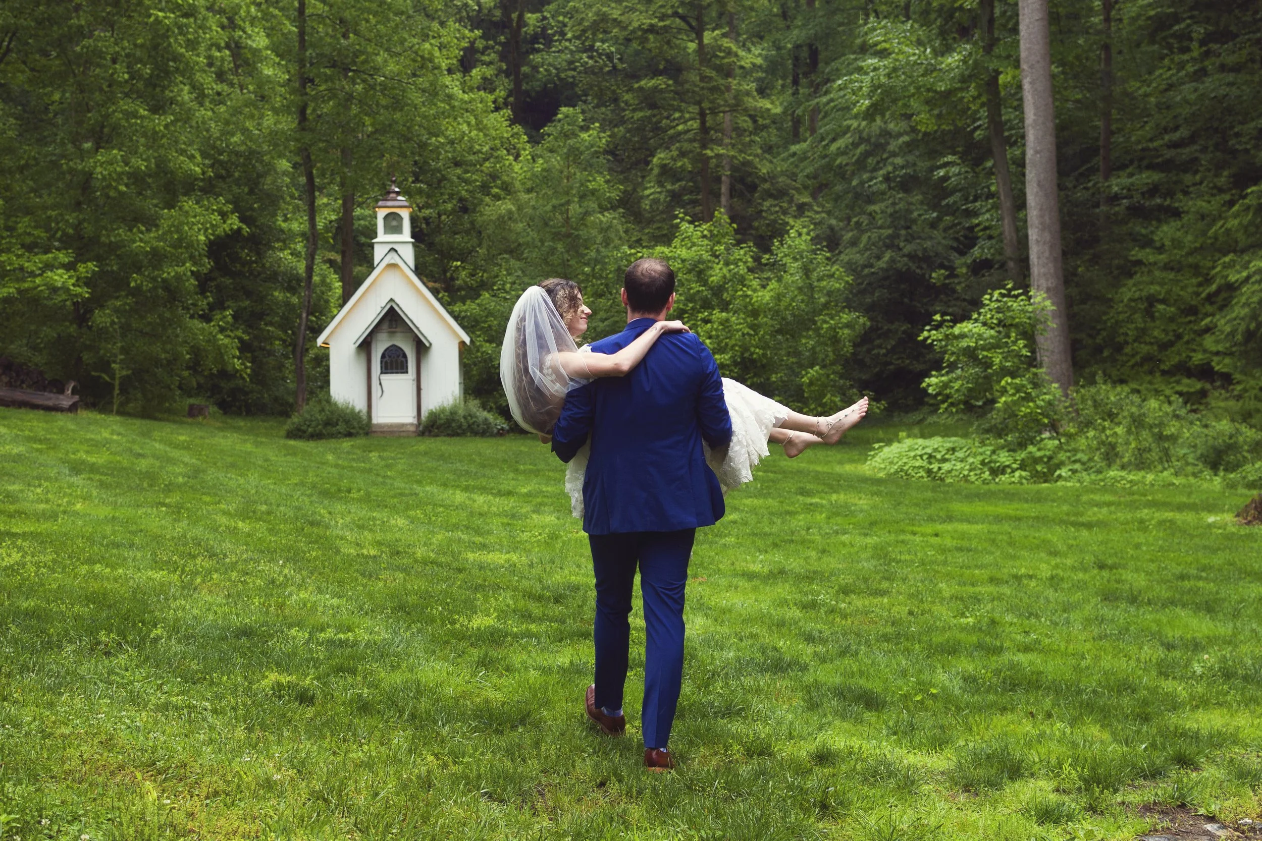 A man in a blue suit carrying a woman in a wedding dress and veil through a lush green garden with a small white chapel in the background.