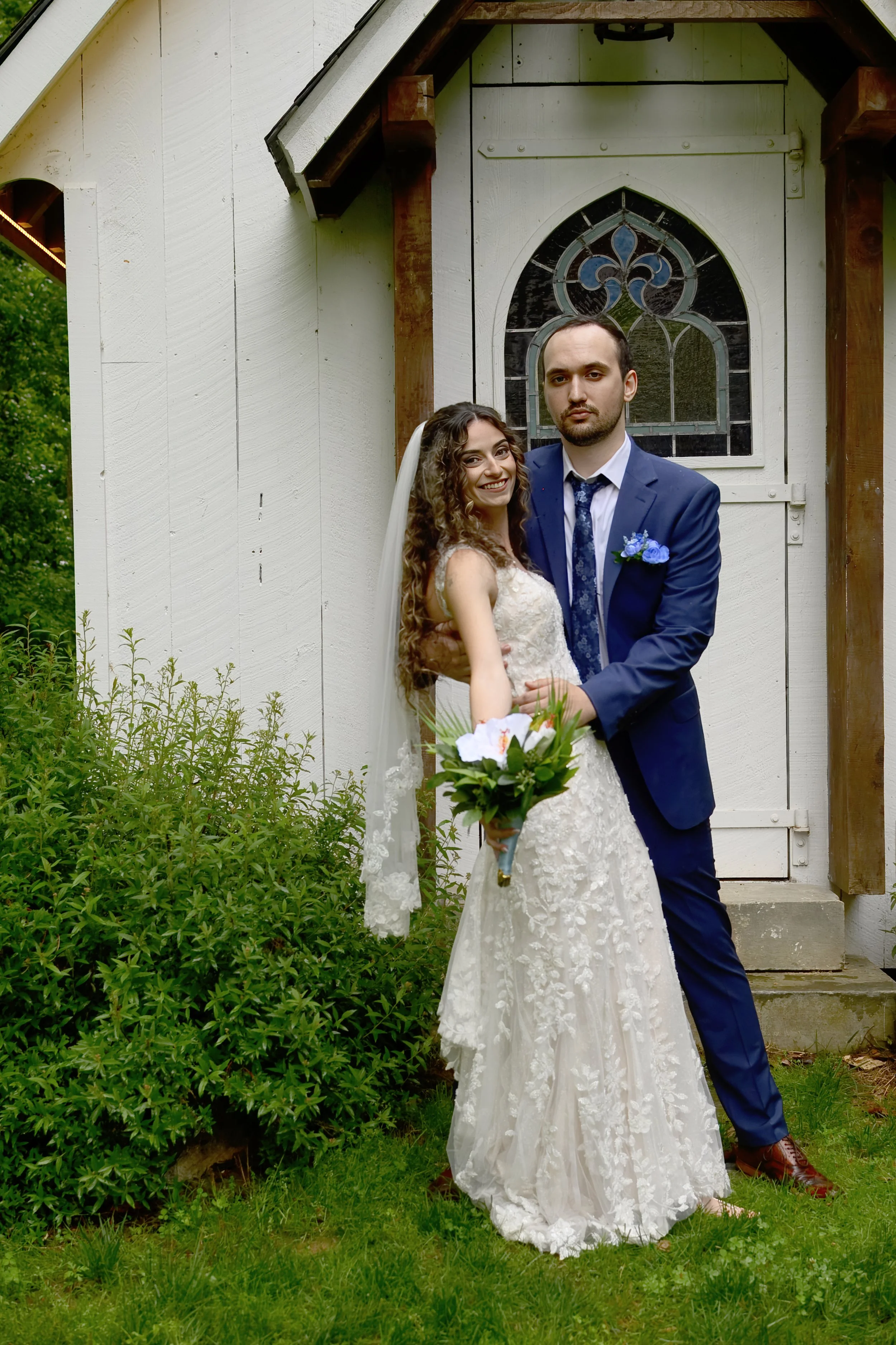 A newlywed couple standing outside near a white wooden building with an arched window. The bride is holding a bouquet of flowers, wearing a lace wedding gown and veil, and smiling at the camera. The groom is dressed in a dark blue suit and tie, with 