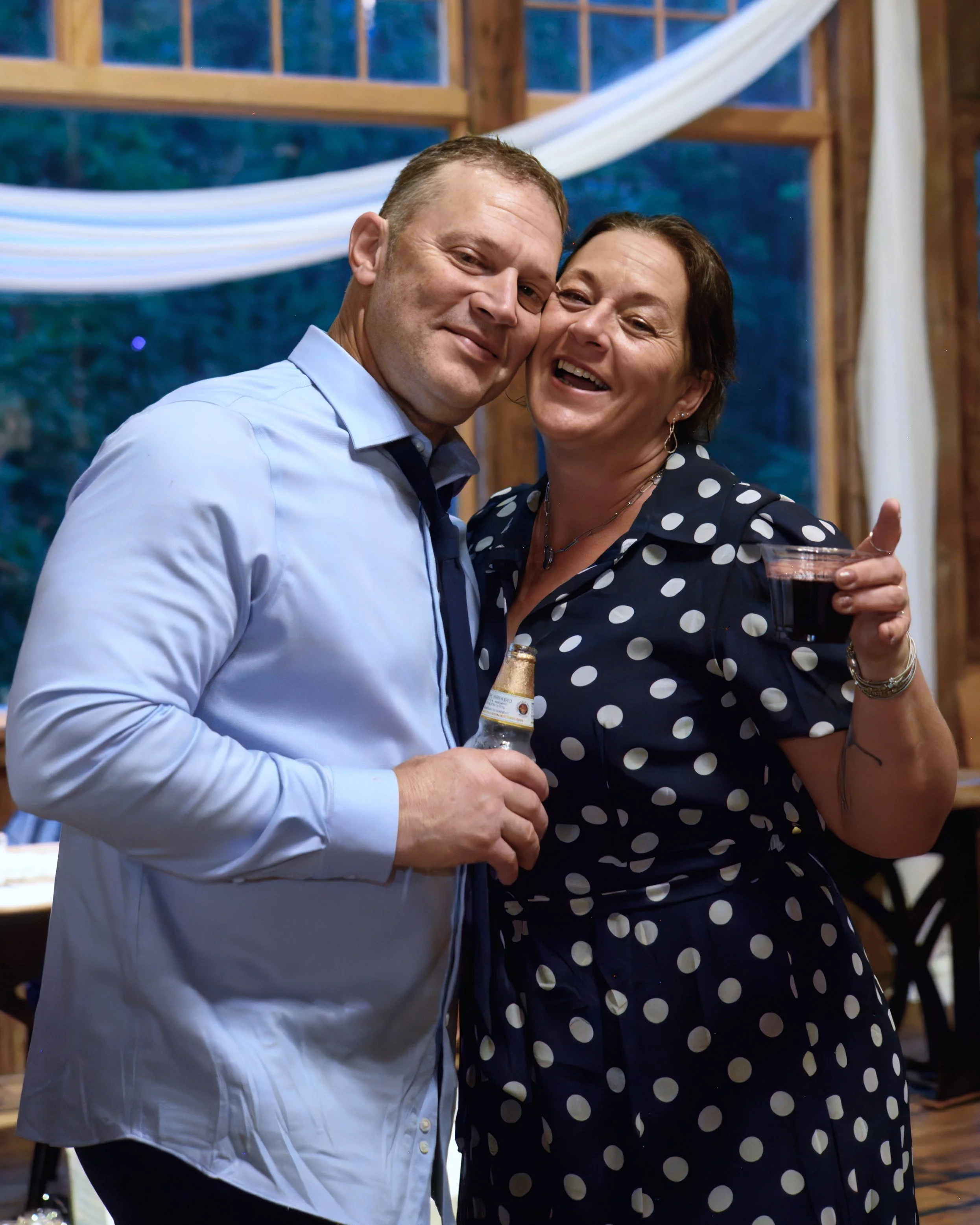 A man in a light blue shirt and a woman in a navy blue polka dot dress are smiling and close together, holding drinks, at a social gathering with a wooden interior and large windows.