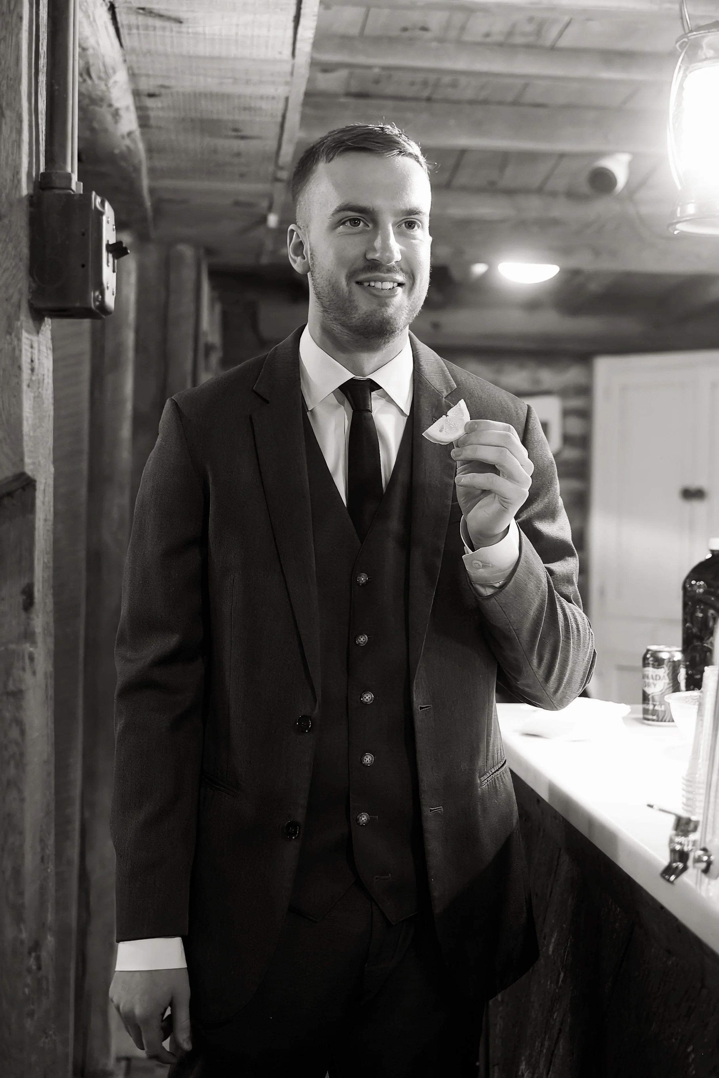 A man in a suit holding a slice of lemon in a rustic indoor setting, illuminated by bright light from the right side.