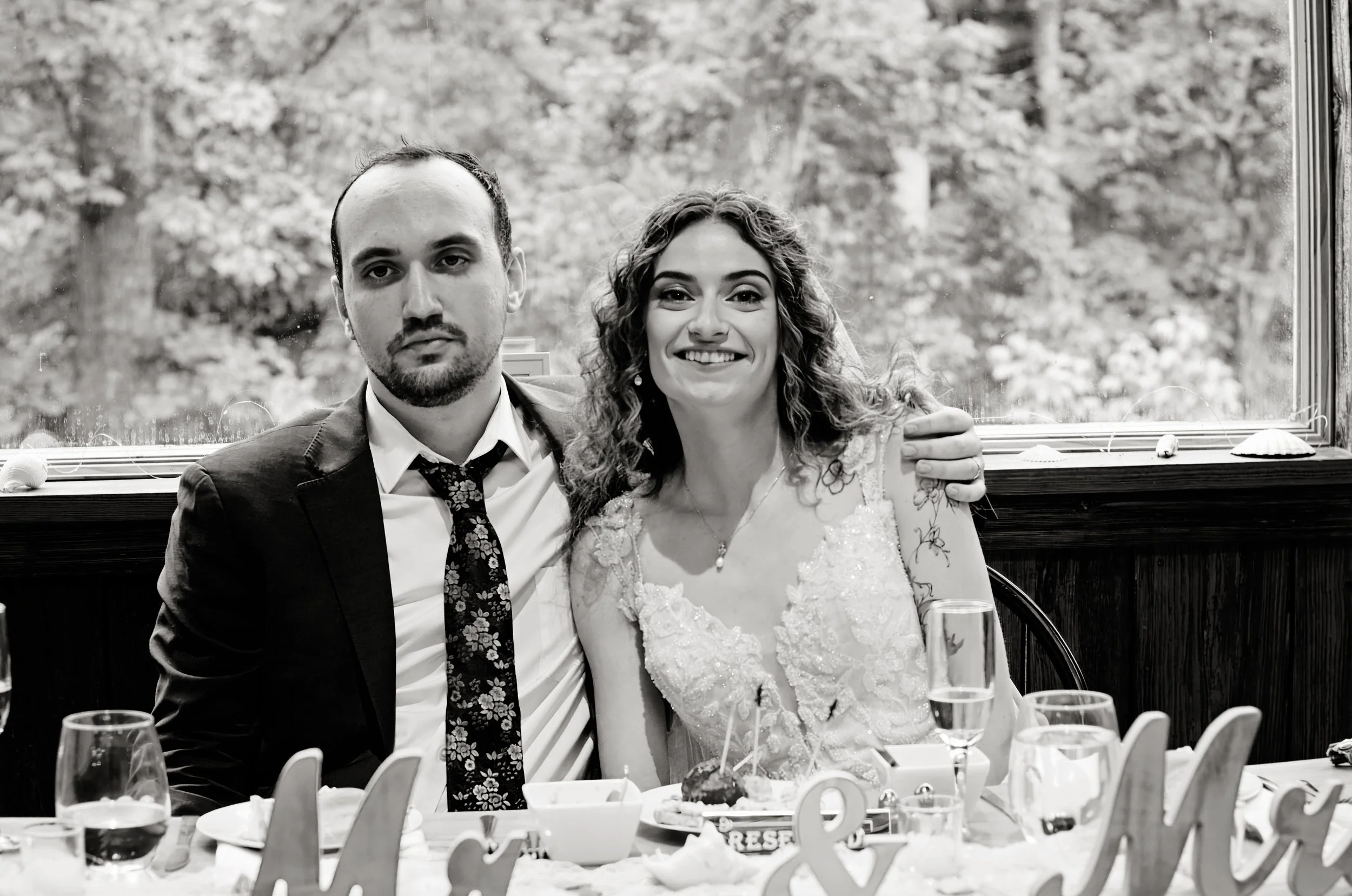 Black and white photo of a couple, likely newlyweds, sitting at a table during a celebration or reception, with decorations and glasses on the table, and outdoors foliage visible through a window behind them.