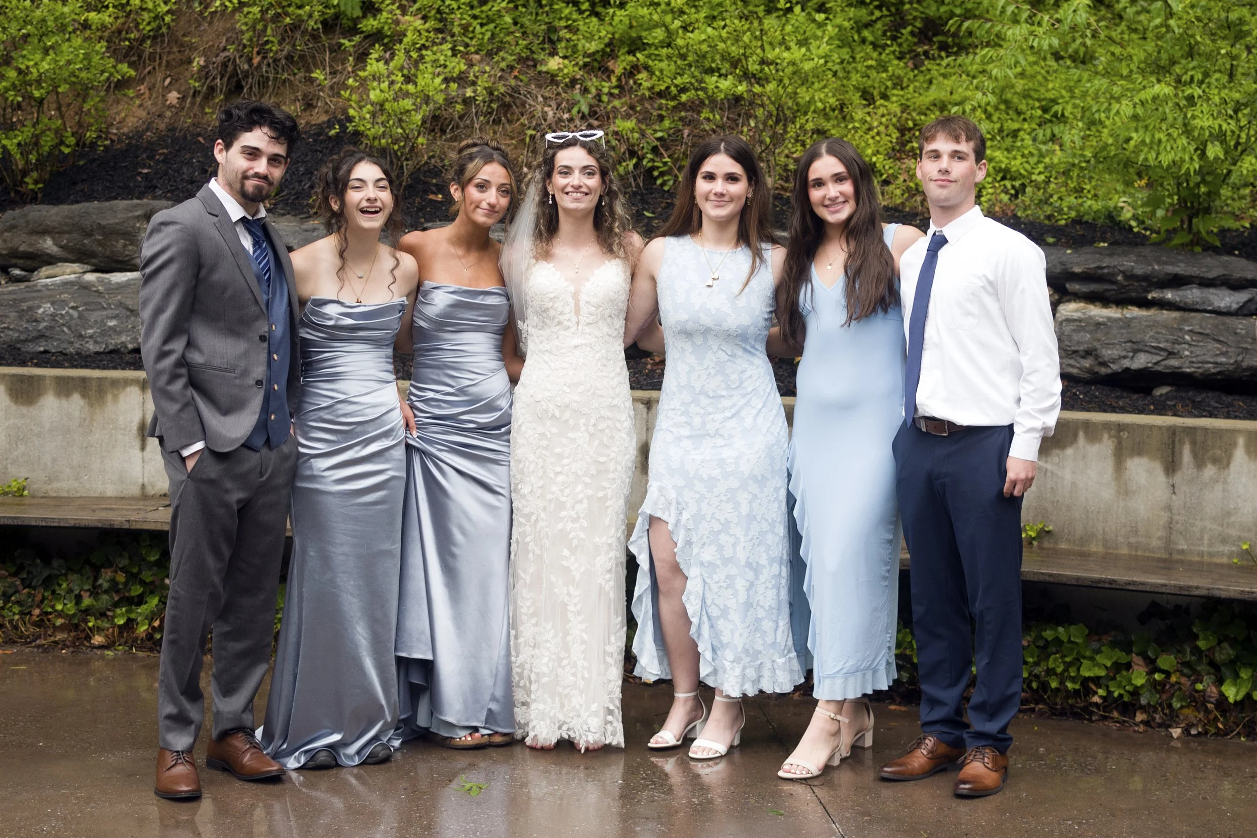 Group of seven young adults dressed in formal attire, standing outdoors with greenery and a rocky background.