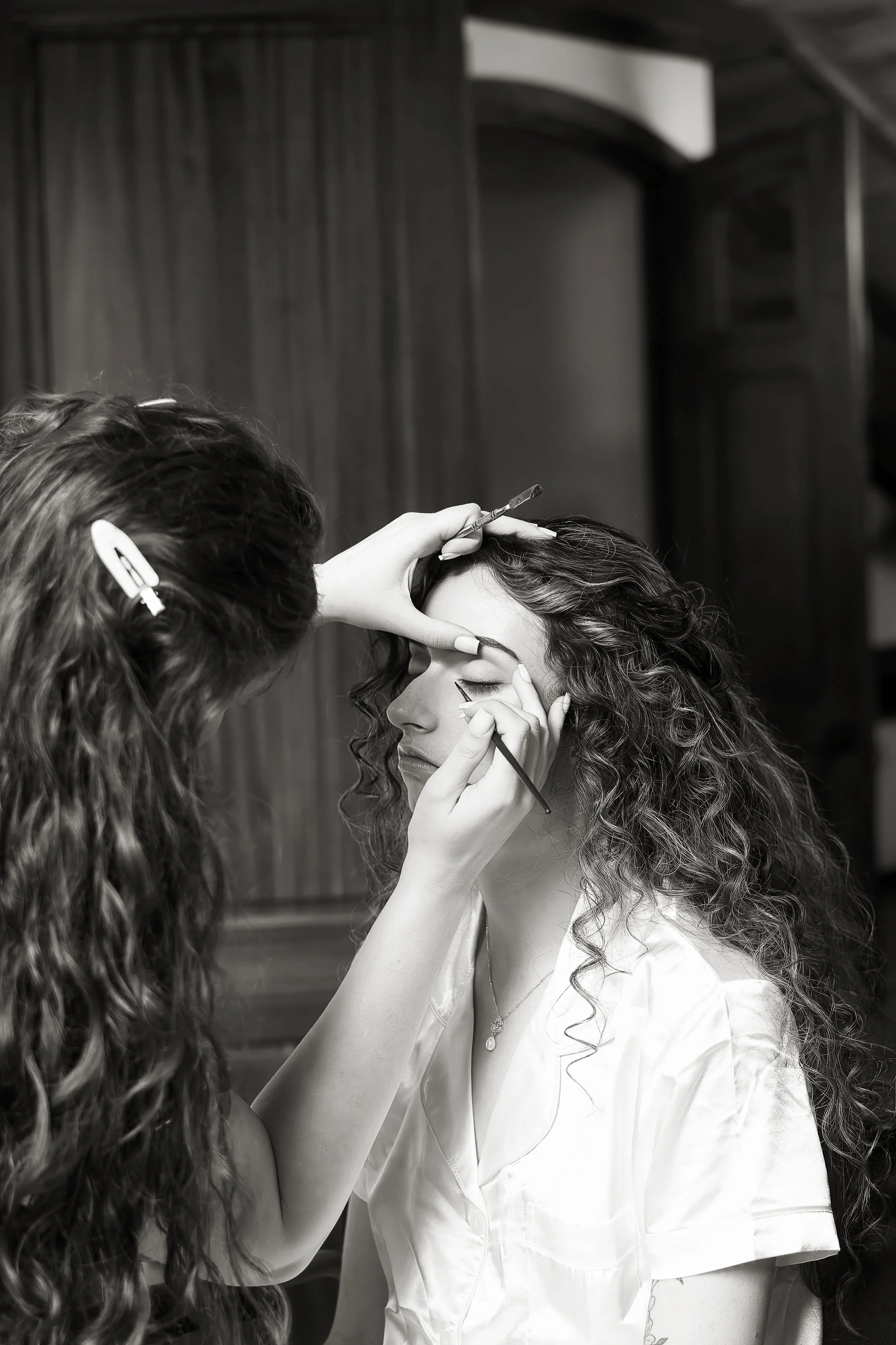 A woman with curly hair being makeuped by another woman with long curly hair, holding a makeup brush and using her hand to steady her face, in a room with wooden walls.