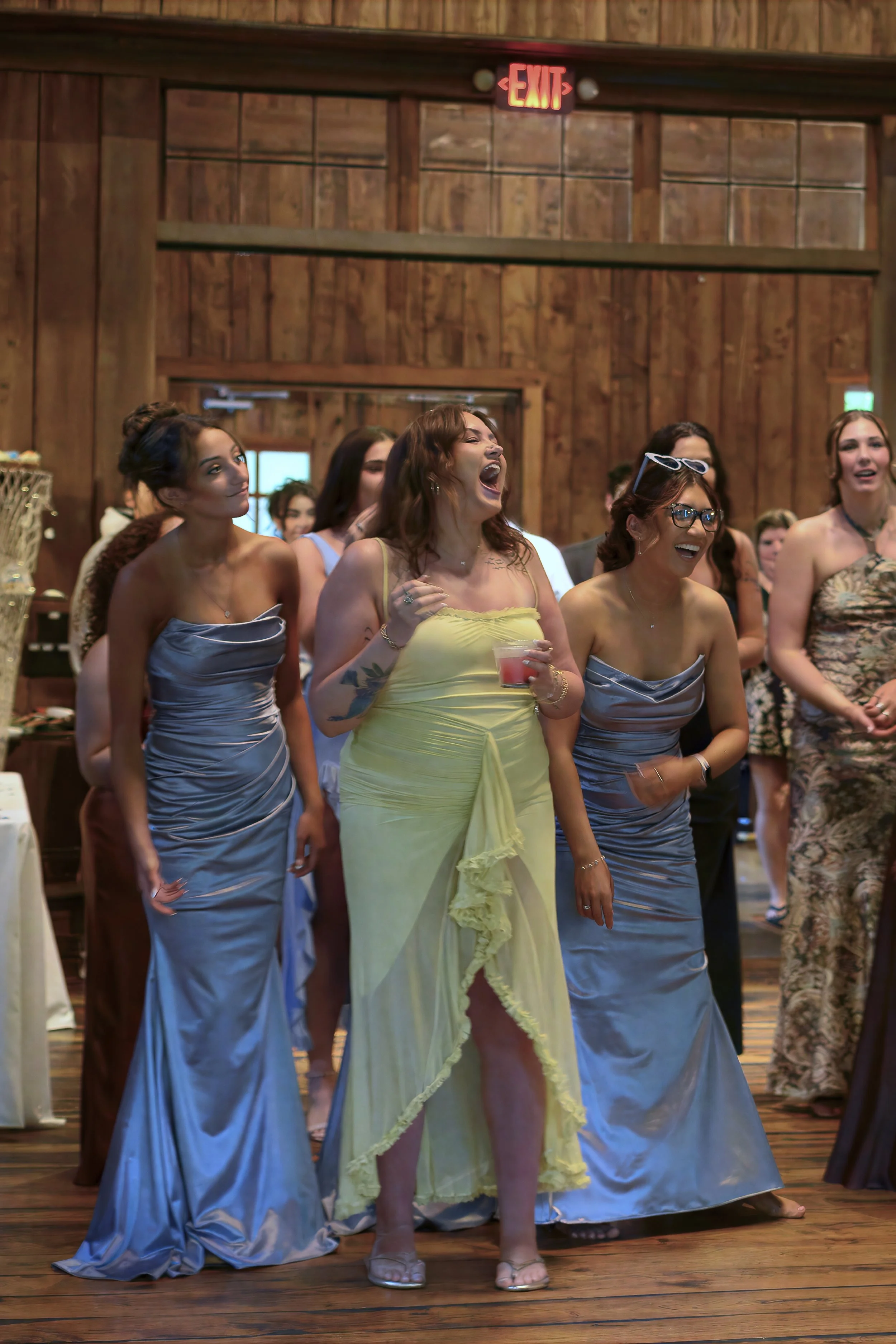 Group of women in formal dresses standing together at what appears to be a wedding reception or celebration inside a rustic wooden venue. The woman in the center is wearing a yellow dress and appears to be laughing or singing. Some women are smiling 