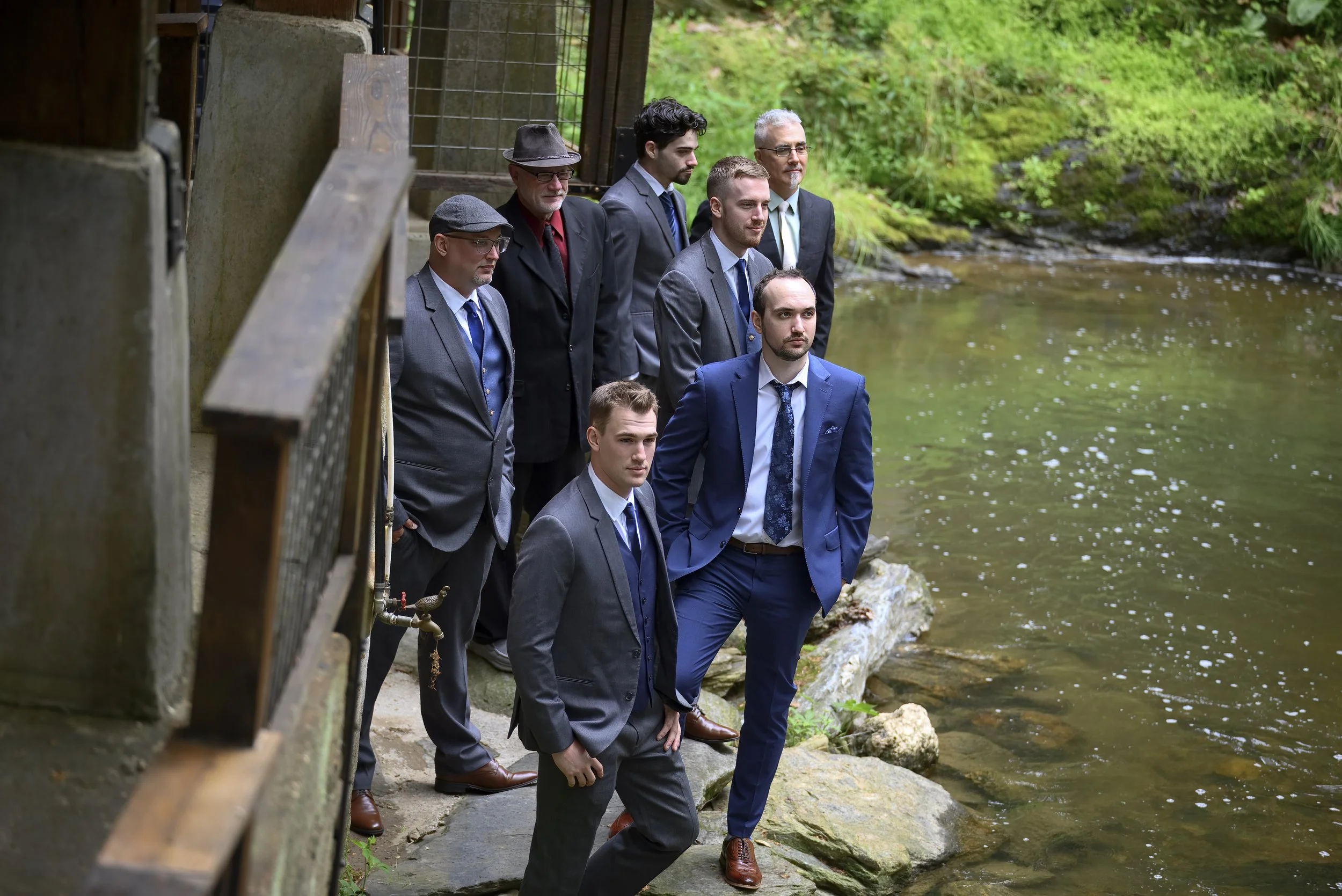 A group of seven men in suits standing by a river, looking at the water, with lush greenery in the background.