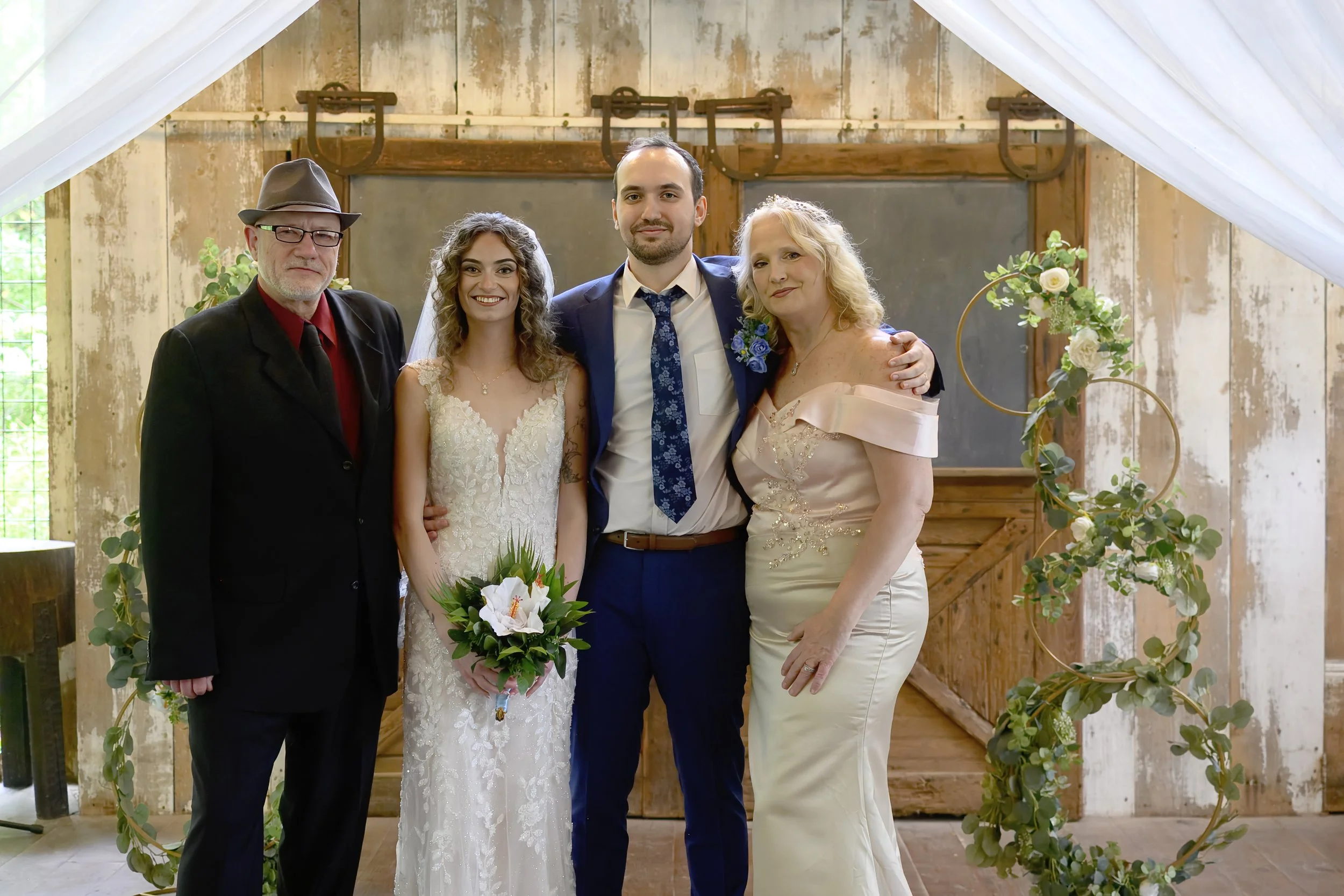A group of five people standing together at a wedding, with a bride in a white lace wedding dress holding a bouquet of white flowers, and the groom in a blue suit. They are flanked by an older man in a black suit and hat, and an older woman in a ligh