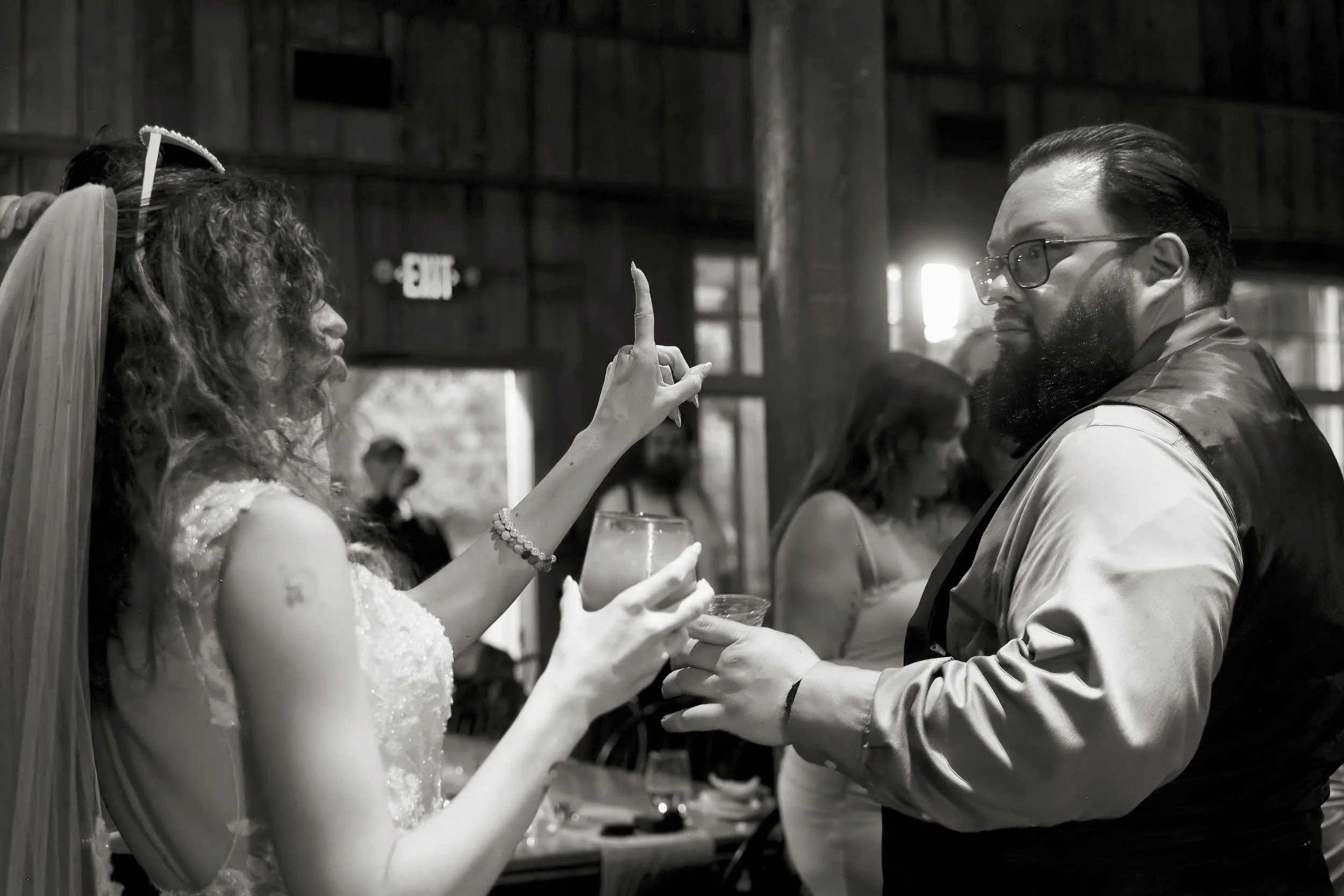 A woman with curly hair, wearing a veil and a wedding dress, raising her finger while talking to a man with glasses and a beard, dressed in a vest, at a wedding reception.