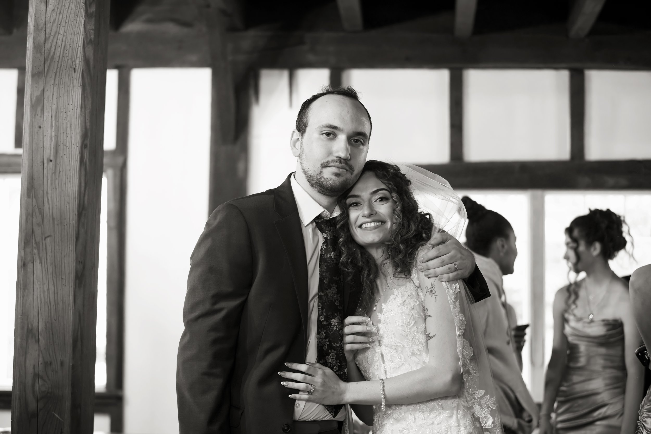A newlywed couple posing for a photo at their wedding reception or ceremony. The groom is wearing a dark suit and the bride is in a white wedding dress with lace details.