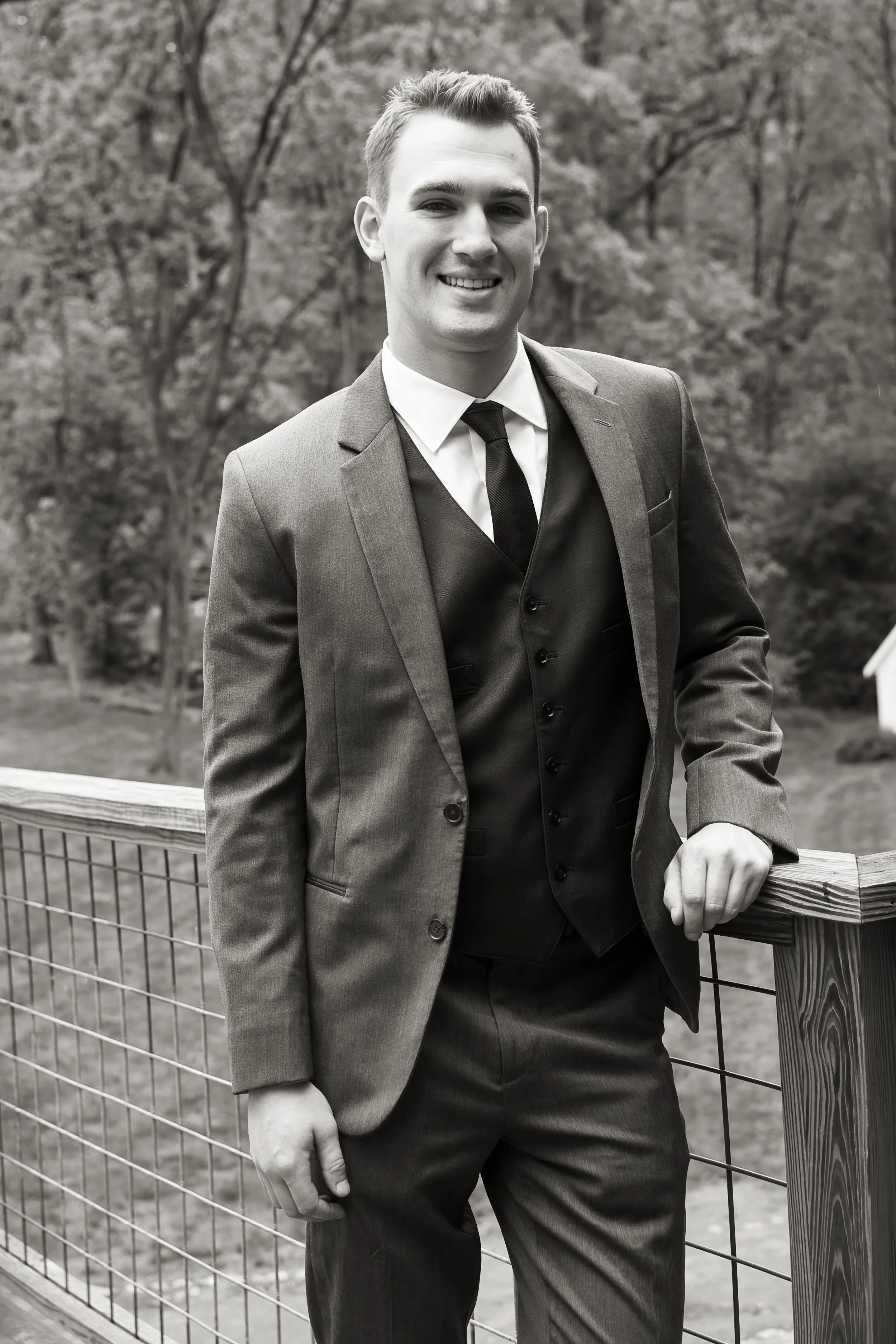 A young man in a suit is leaning on a wooden and wire fence outdoors in a wooded area, smiling at the camera.