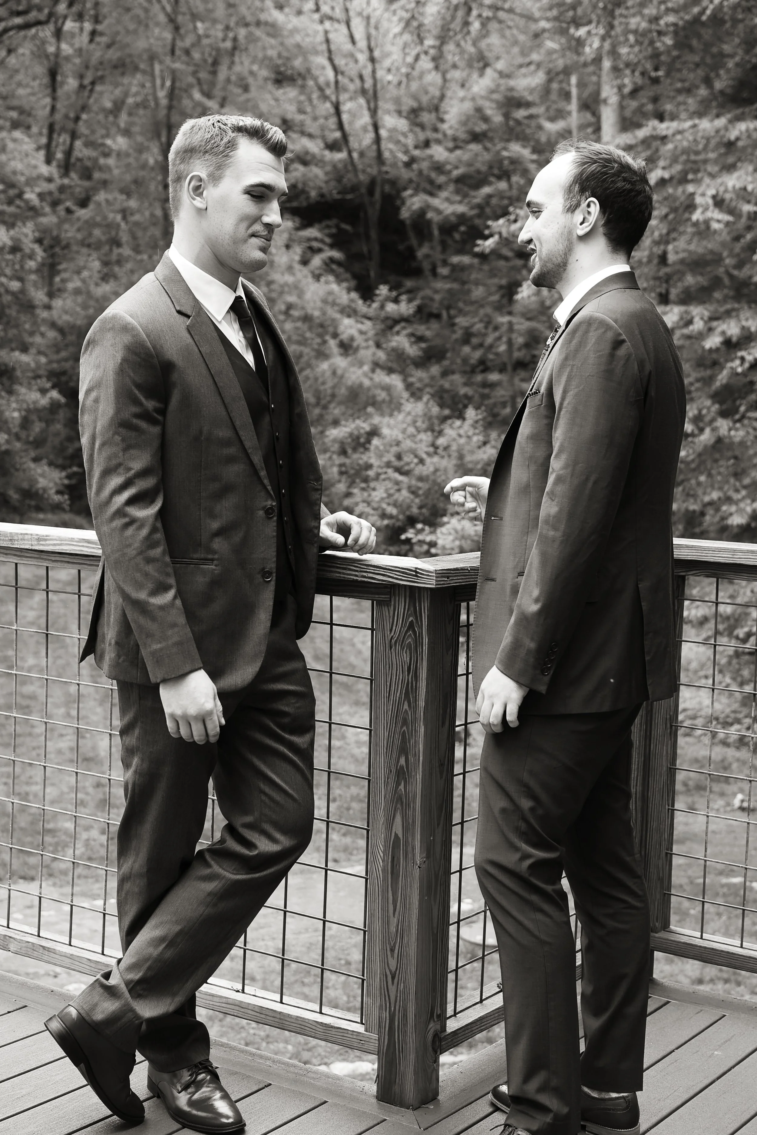 Two men in formal suits stand on a wooden balcony with a tree-lined background, engaging in conversation.