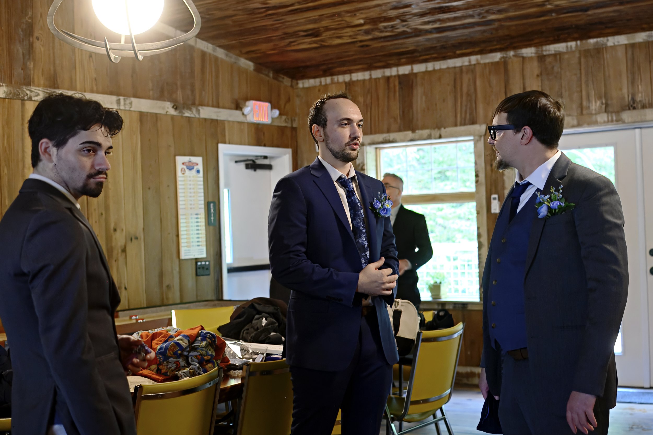 Three men in suits with boutonnières talking indoors with wooden walls and partially visible items on a table.