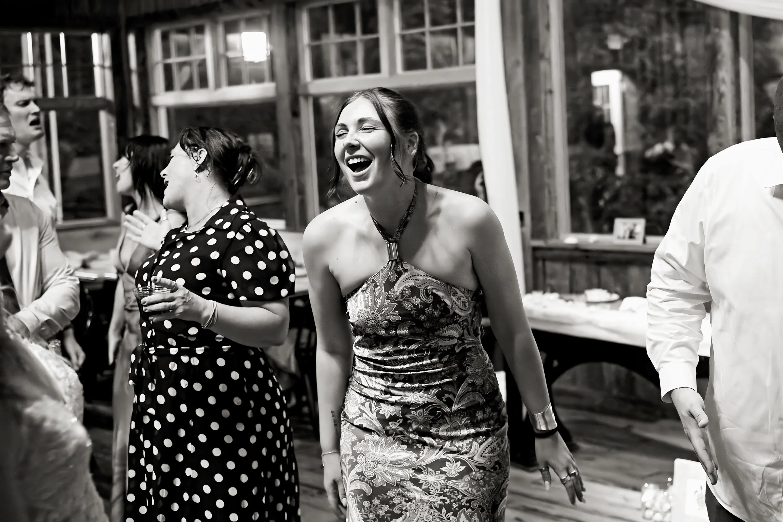 People at a social gathering, with a woman in the center laughing joyfully, dressed in a patterned dress, holding a drink, in an open, rustic setting with wooden beams and windows.