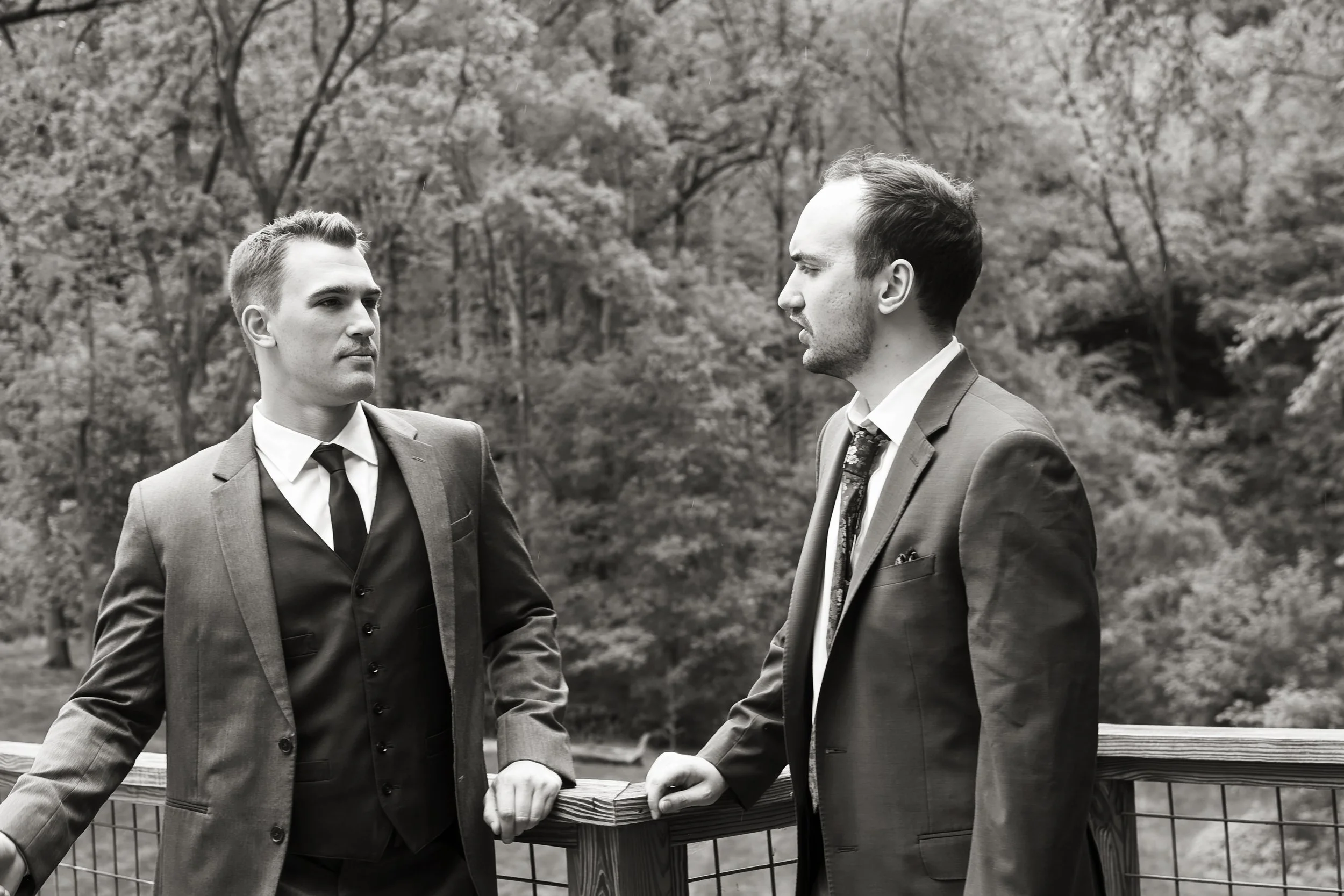 Two men in suits engaged in conversation outdoors, standing near a wooden bench and a fence, with trees in the background.