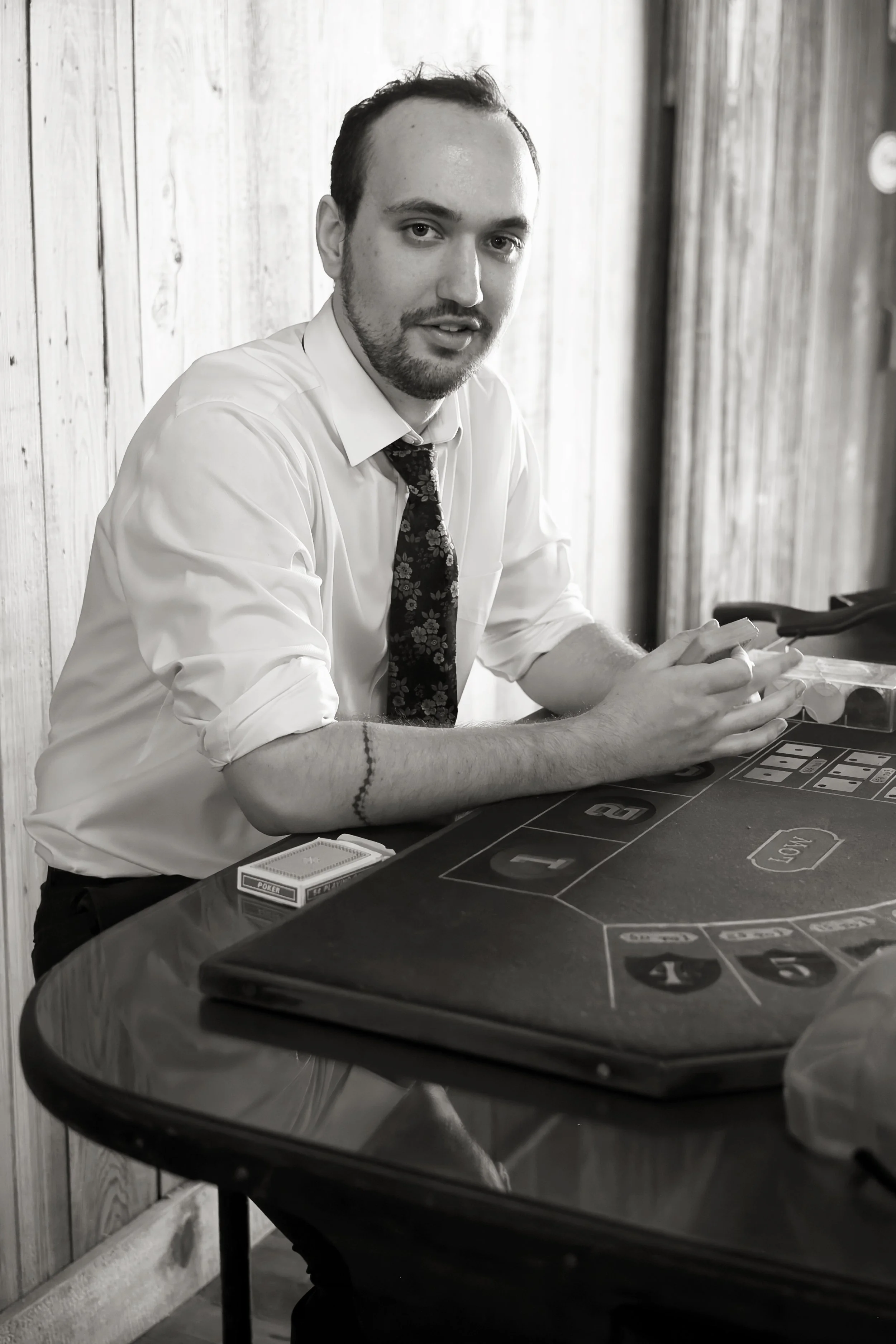 Man with a beard, wearing a white shirt and floral tie, sitting at a blackjack table with a deck of playing cards and poker chips, on a wooden wall background.