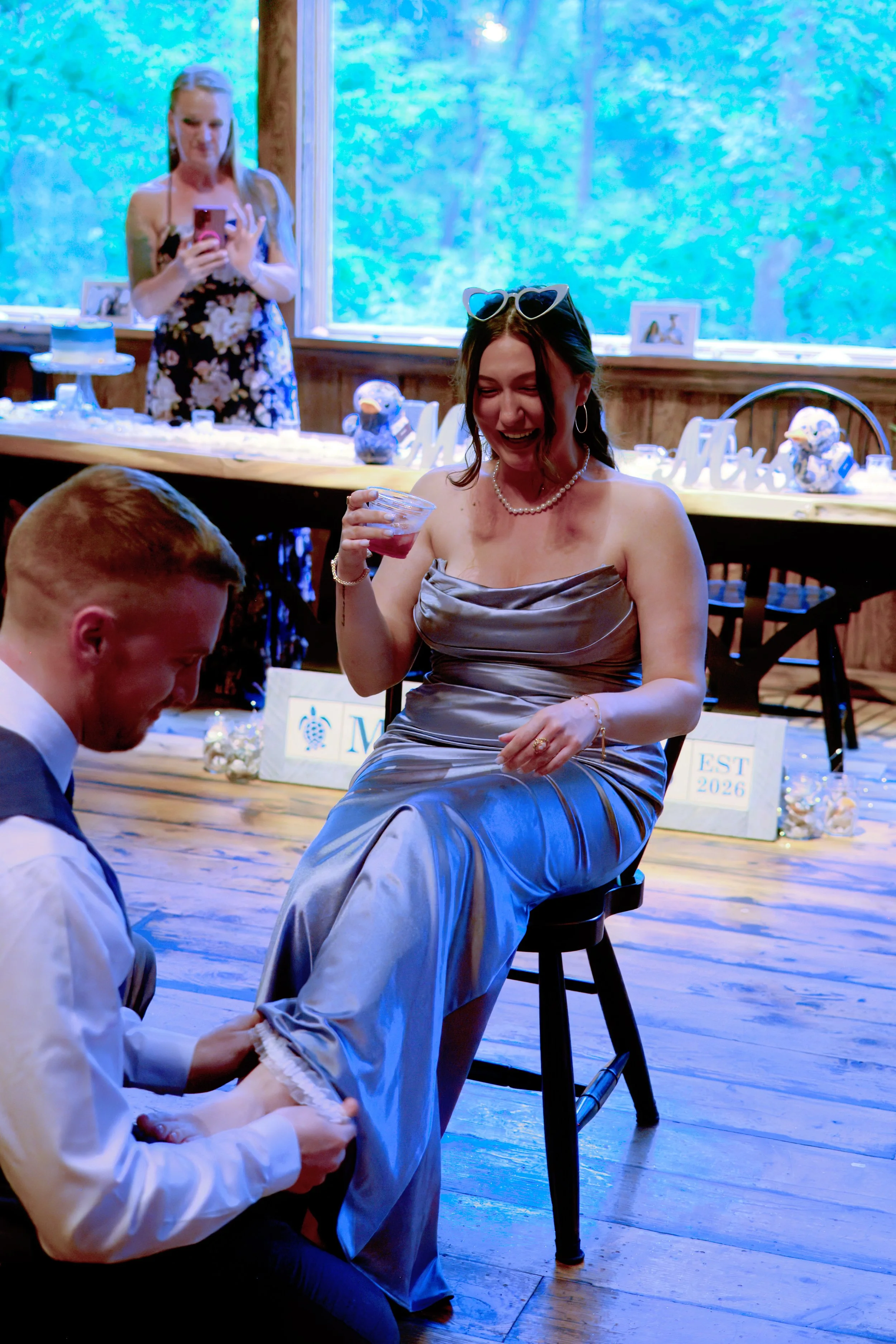 A woman in a strapless blue dress with heart-shaped sunglasses on her head is smiling and holding a drink. There is a man kneeling in front of her, holding her foot. In the background, a woman is taking a photo. The setting appears to be a celebratio