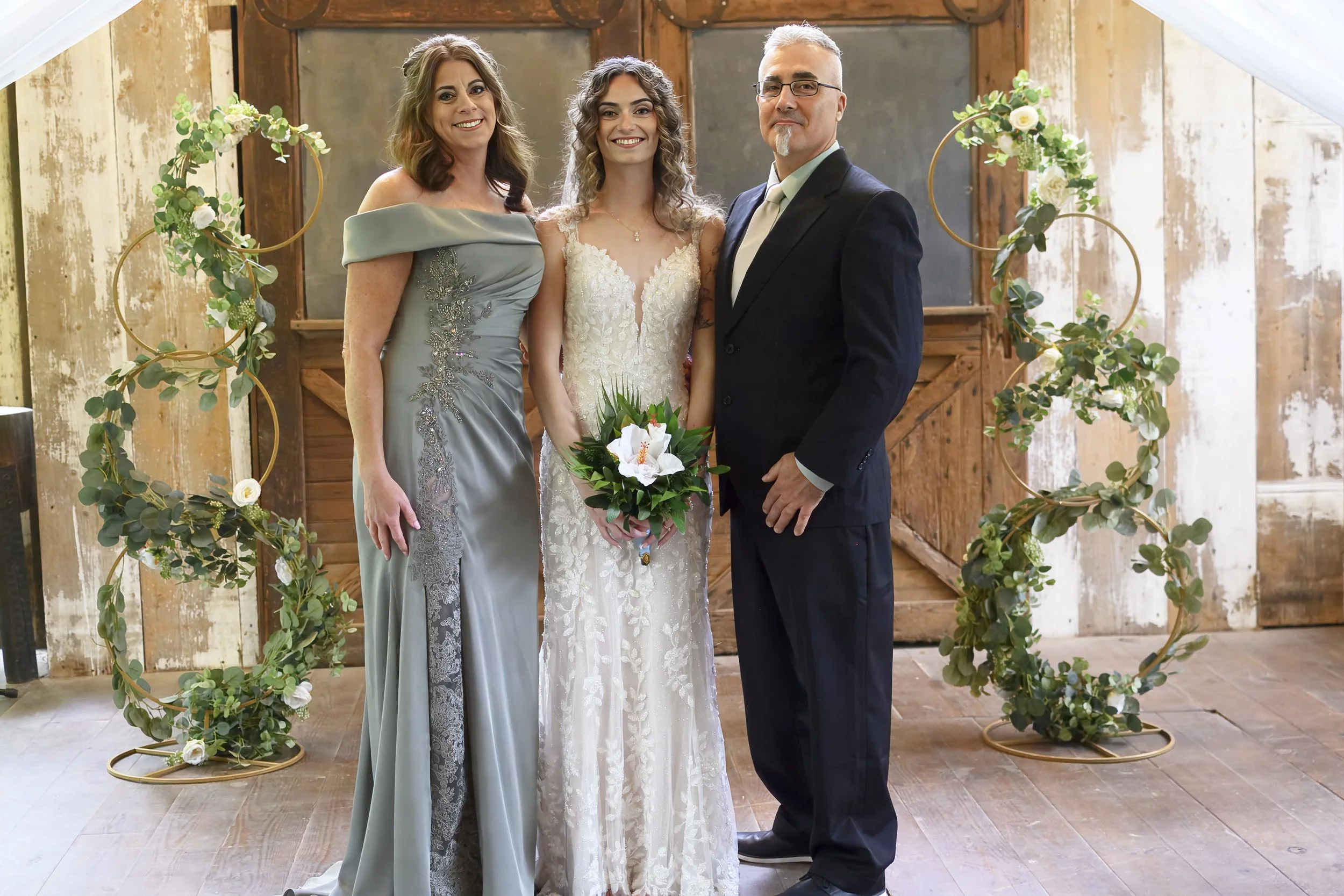 Three people standing together at a wedding ceremony, with a bride in the middle holding a bouquet, flanked by a woman in a gray off-shoulder dress and a man in a black suit, in front of a circular floral arch.