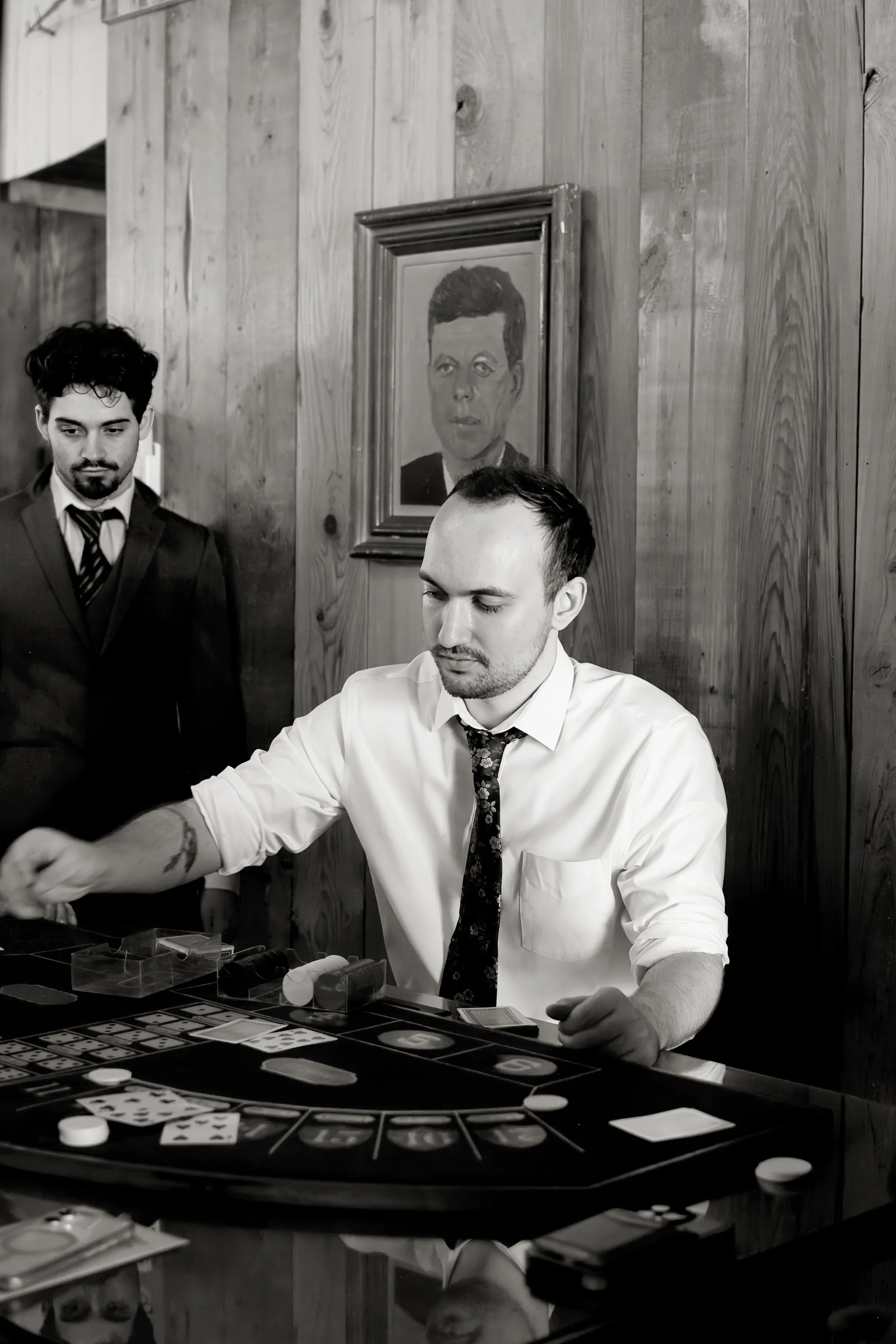 Two men playing blackjack at a casino table, with cards and chips visible, while a framed portrait hangs on a wooden wall behind them.