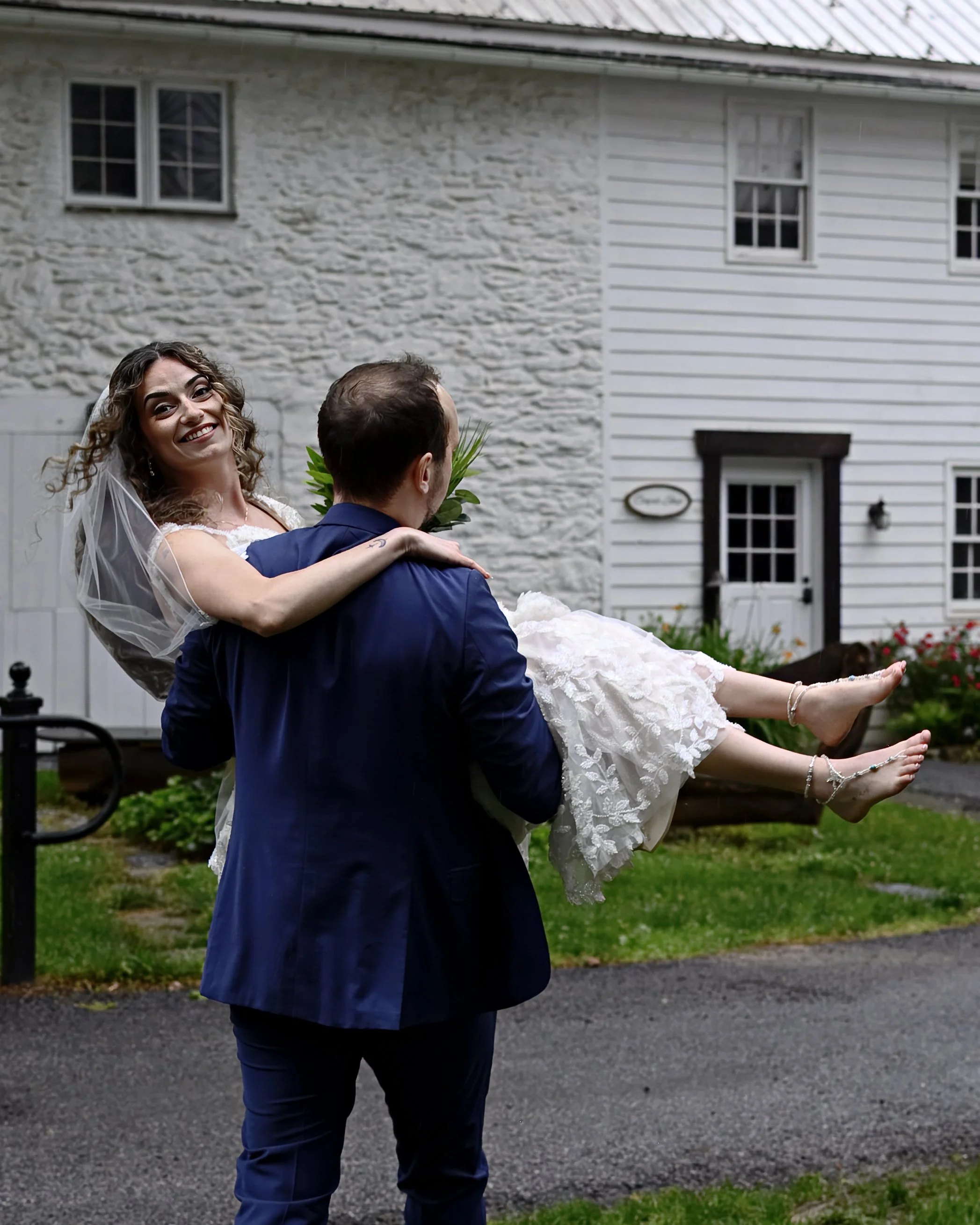 A man in a blue suit holding a smiling woman in a wedding dress with a veil, outdoors in front of a white house with windows and a door, on a cloudy day.